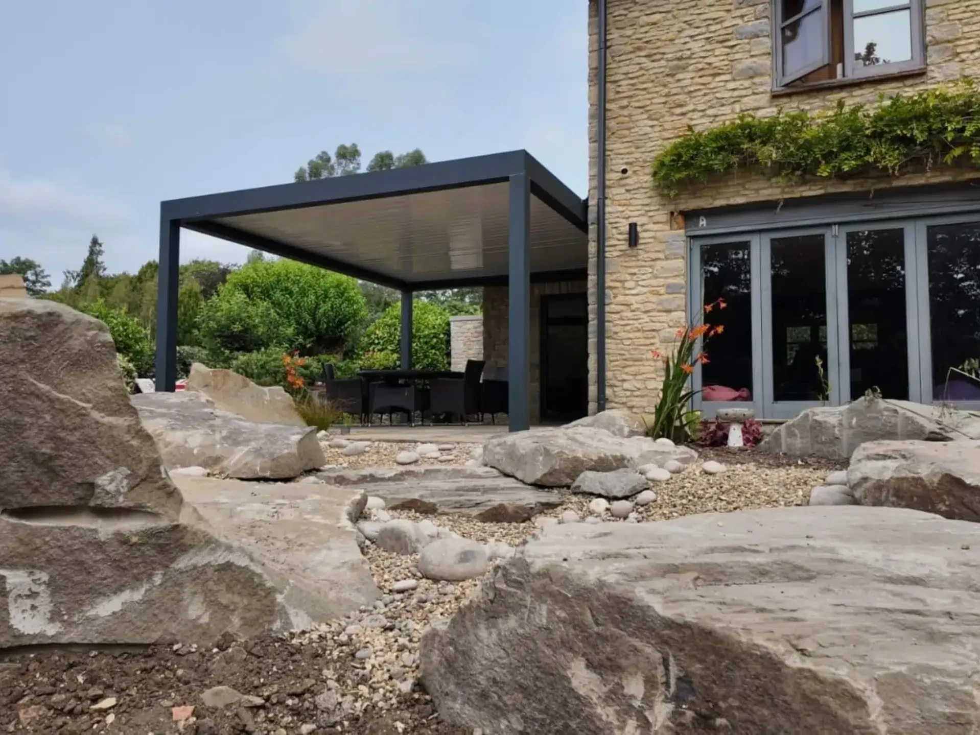 Anthracite grey aluminium frame pergola with white aluminium bioclimatic roof slats, furnished with dark brown rattan garden chairs and a glass table.