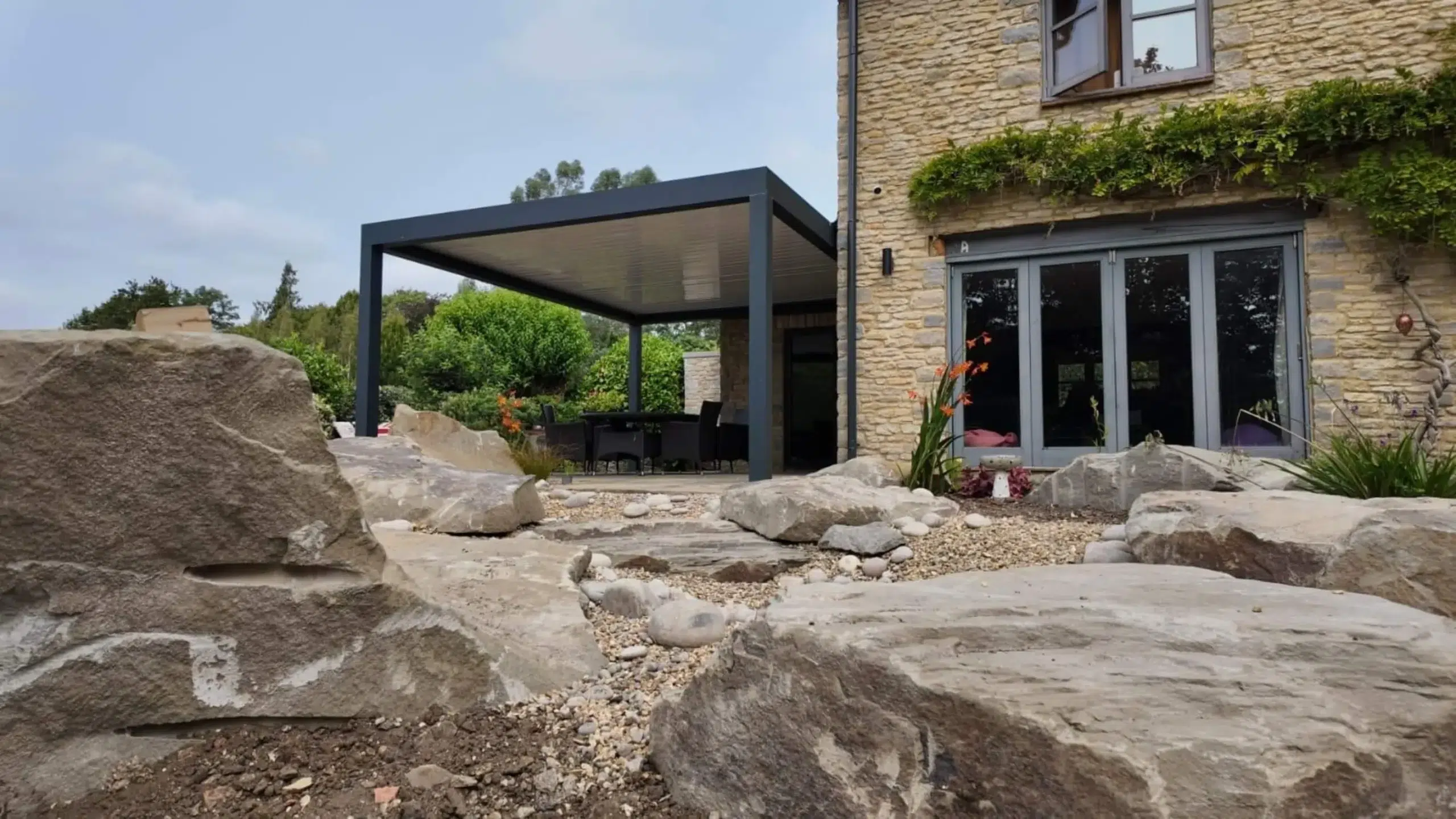 Anthracite grey aluminium frame pergola with white aluminium bioclimatic roof slats, furnished with dark brown rattan garden chairs and a glass table.