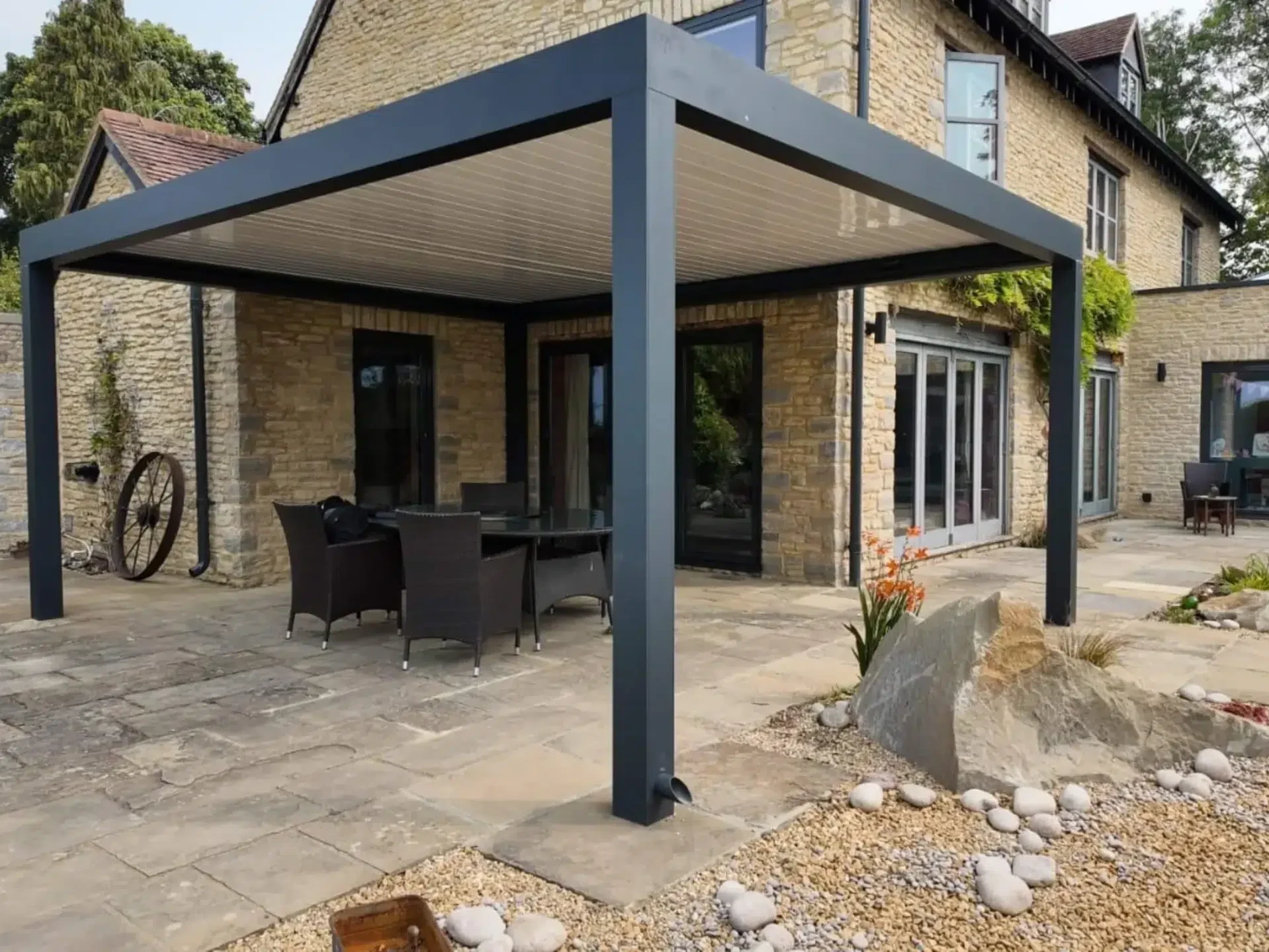 Anthracite grey aluminium frame pergola with white aluminium bioclimatic roof slats, furnished with dark brown rattan garden chairs and a glass table.