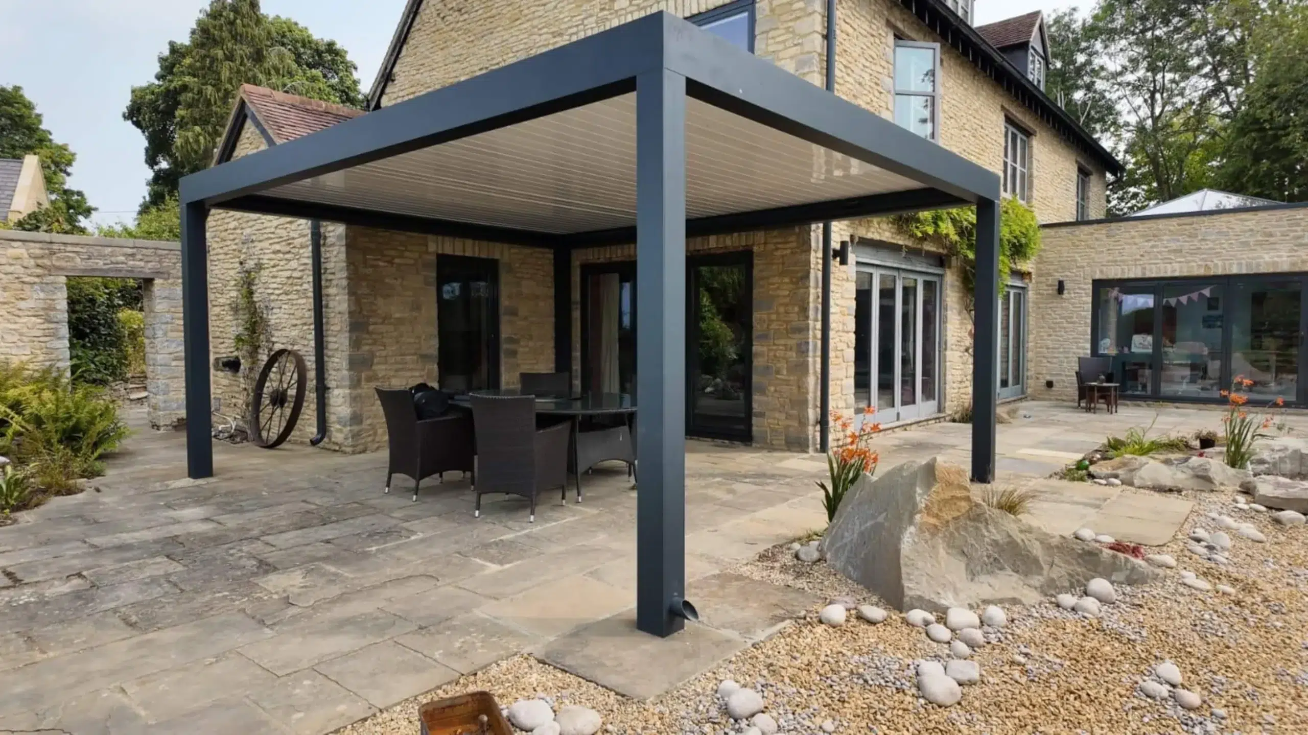 Anthracite grey aluminium frame pergola with white aluminium bioclimatic roof slats, furnished with dark brown rattan garden chairs and a glass table.