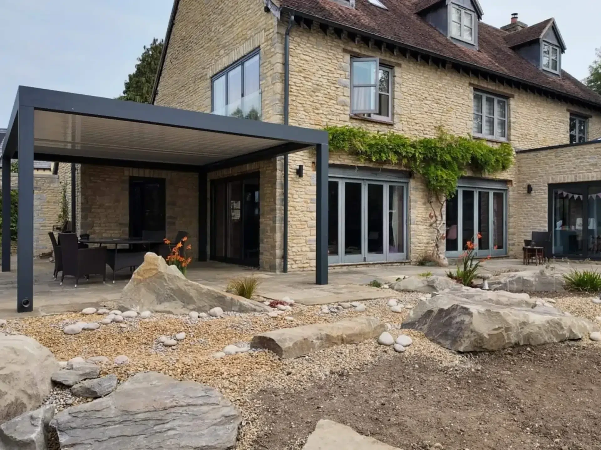 Anthracite grey aluminium frame pergola with white aluminium bioclimatic roof slats, furnished with dark brown rattan garden chairs and a glass table.