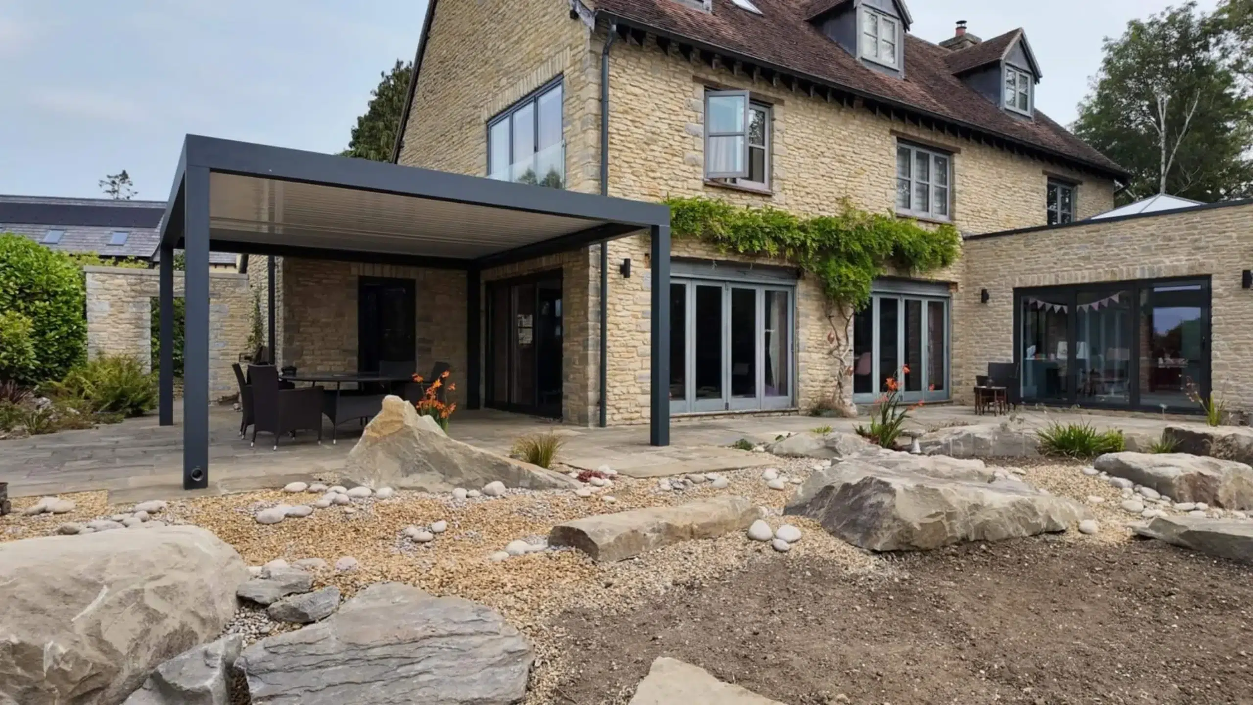 Anthracite grey aluminium frame pergola with white aluminium bioclimatic roof slats, furnished with dark brown rattan garden chairs and a glass table.