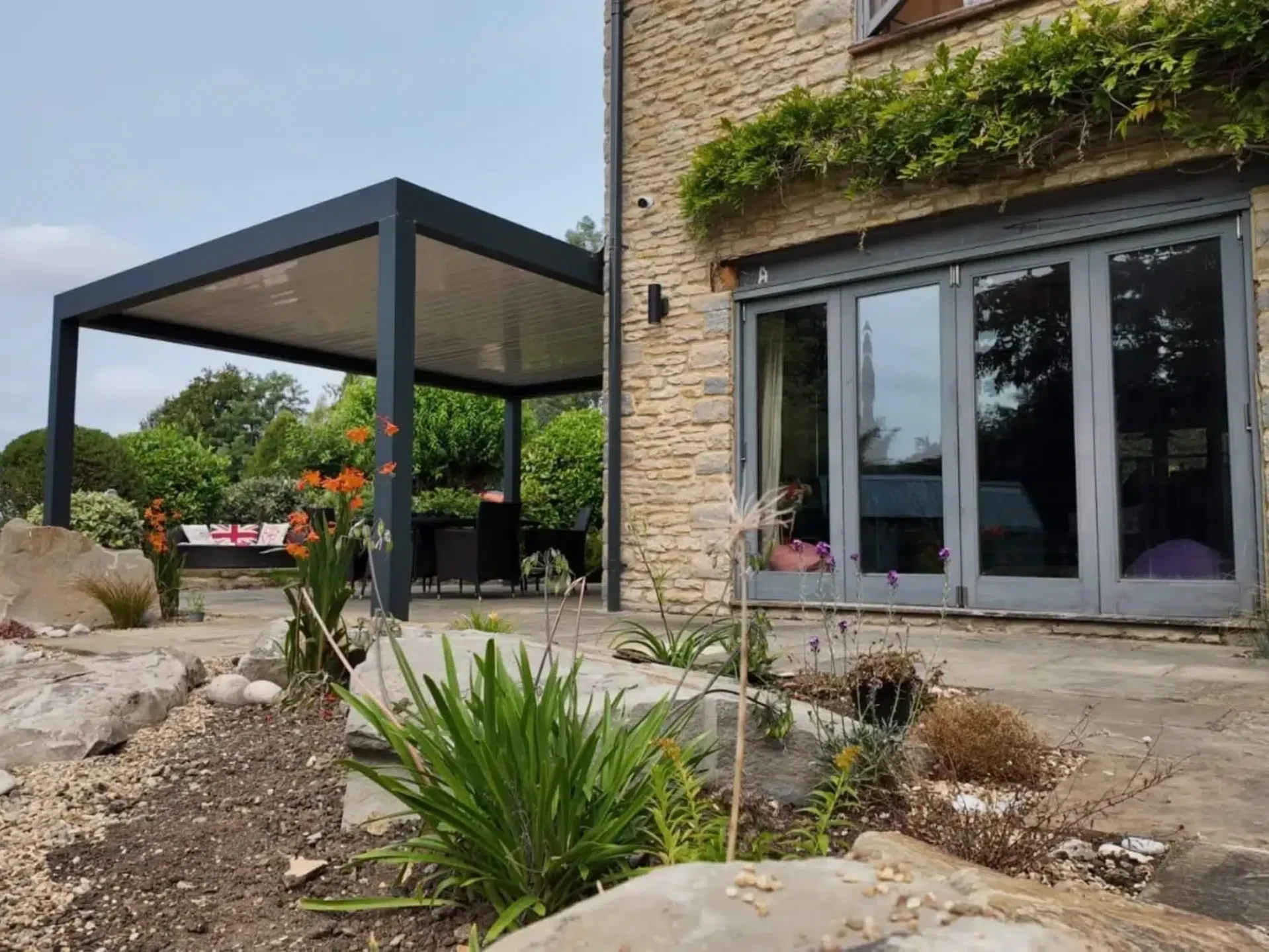 Anthracite grey aluminium frame pergola with white aluminium bioclimatic roof slats, furnished with dark brown rattan garden chairs and a glass table.