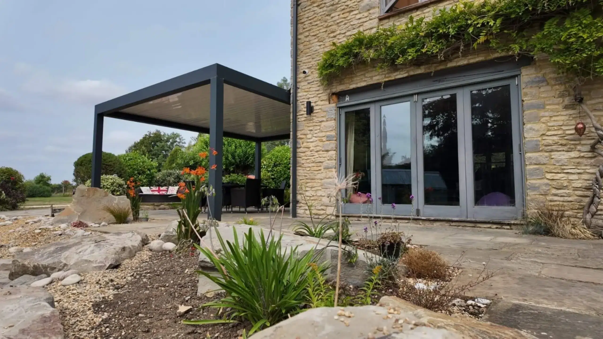 Anthracite grey aluminium frame pergola with white aluminium bioclimatic roof slats, furnished with dark brown rattan garden chairs and a glass table.