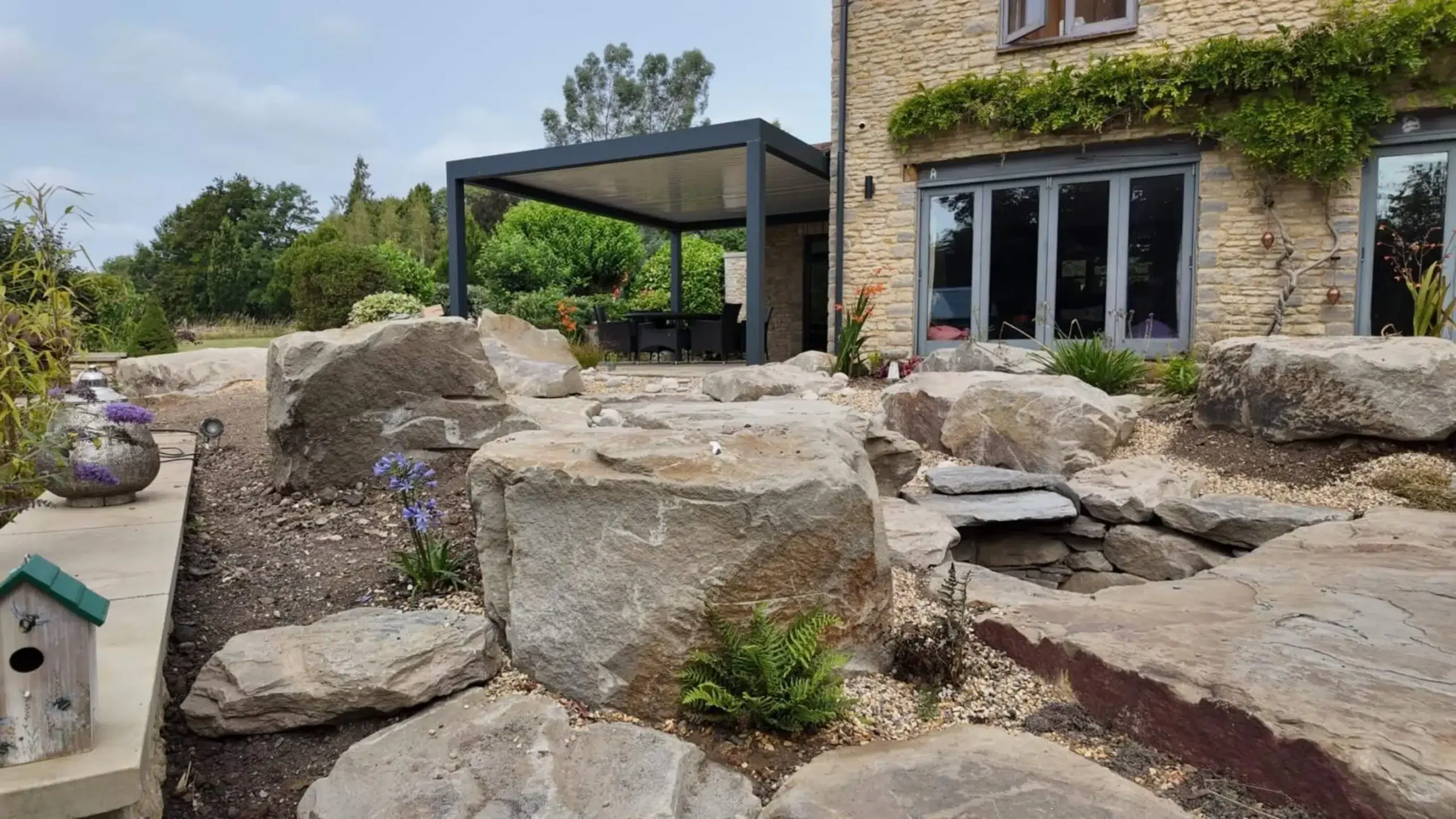 Anthracite grey aluminium frame pergola with white aluminium bioclimatic roof slats, furnished with dark brown rattan garden chairs and a glass table.