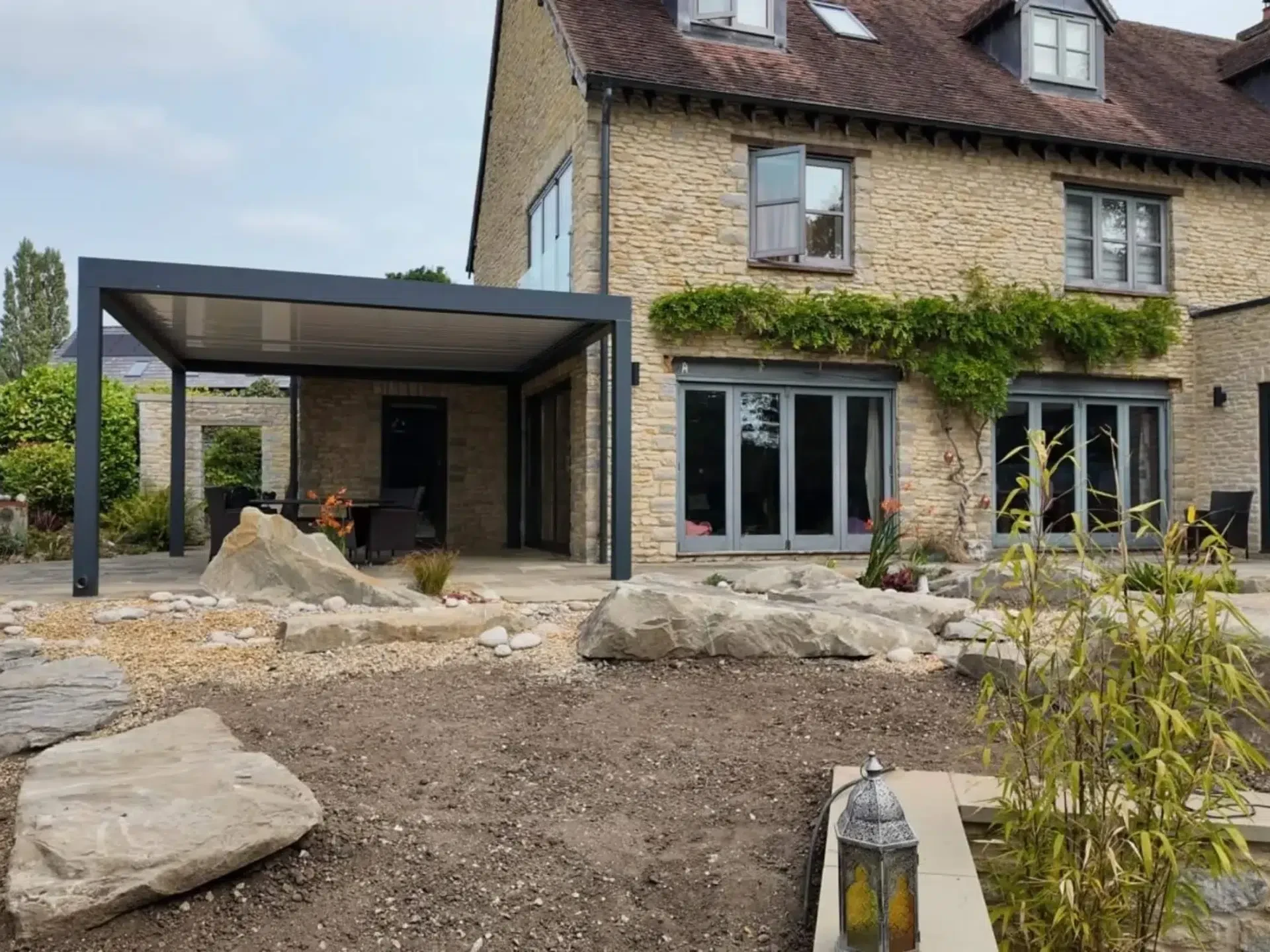 Anthracite grey aluminium frame pergola with white aluminium bioclimatic roof slats, furnished with dark brown rattan garden chairs and a glass table.