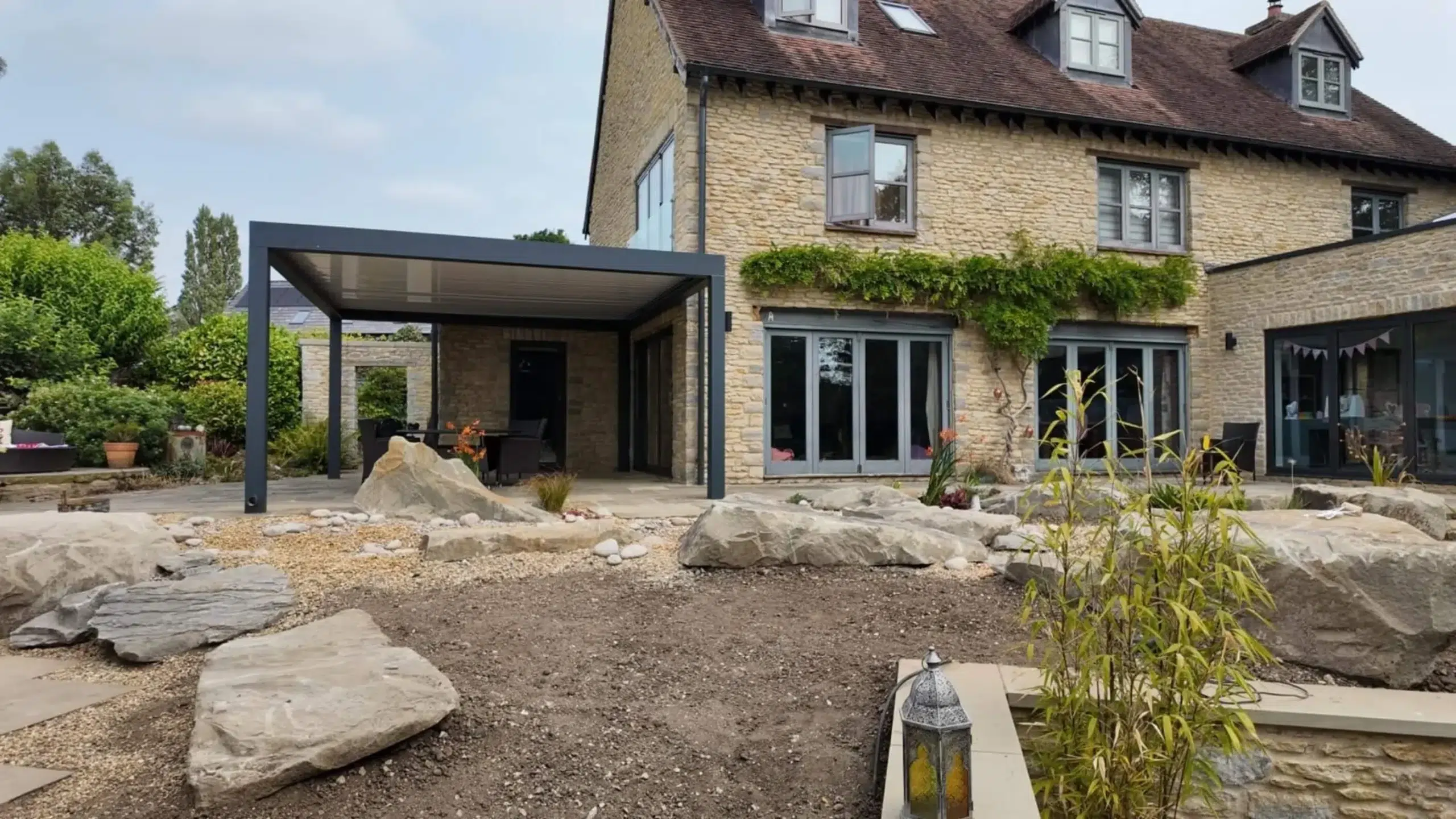 Anthracite grey aluminium frame pergola with white aluminium bioclimatic roof slats, furnished with dark brown rattan garden chairs and a glass table.