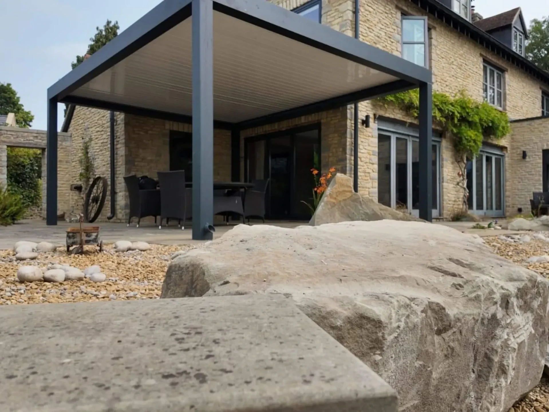 Anthracite grey aluminium frame pergola with white aluminium bioclimatic roof slats, furnished with dark brown rattan garden chairs and a glass table.