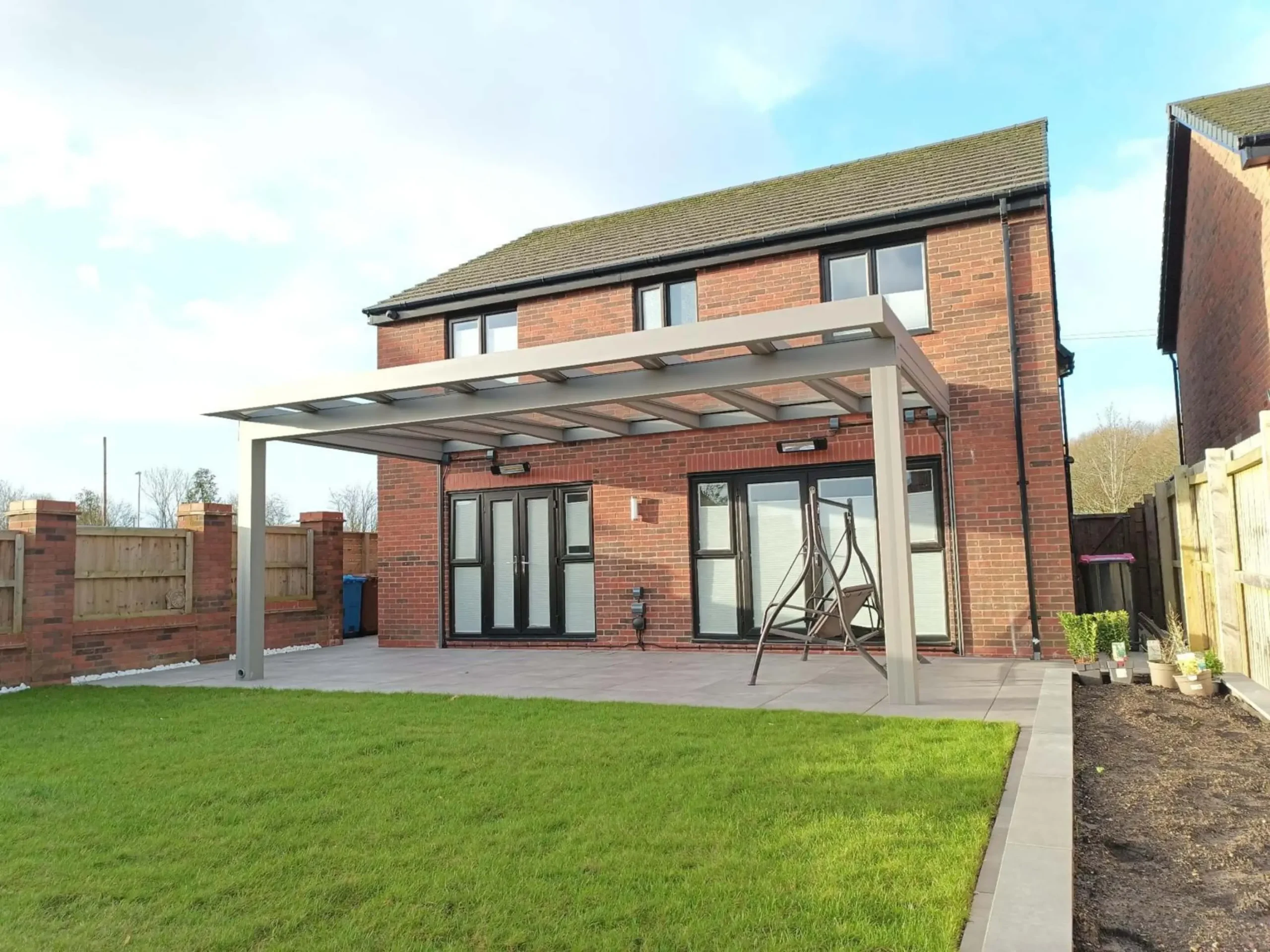 A silver grey aluminium cube line veranda with a laminated glass roof, remote-controlled side screens and black wall-mounted infrared heaters, furnished with a dark grey 3 seater swing chair.