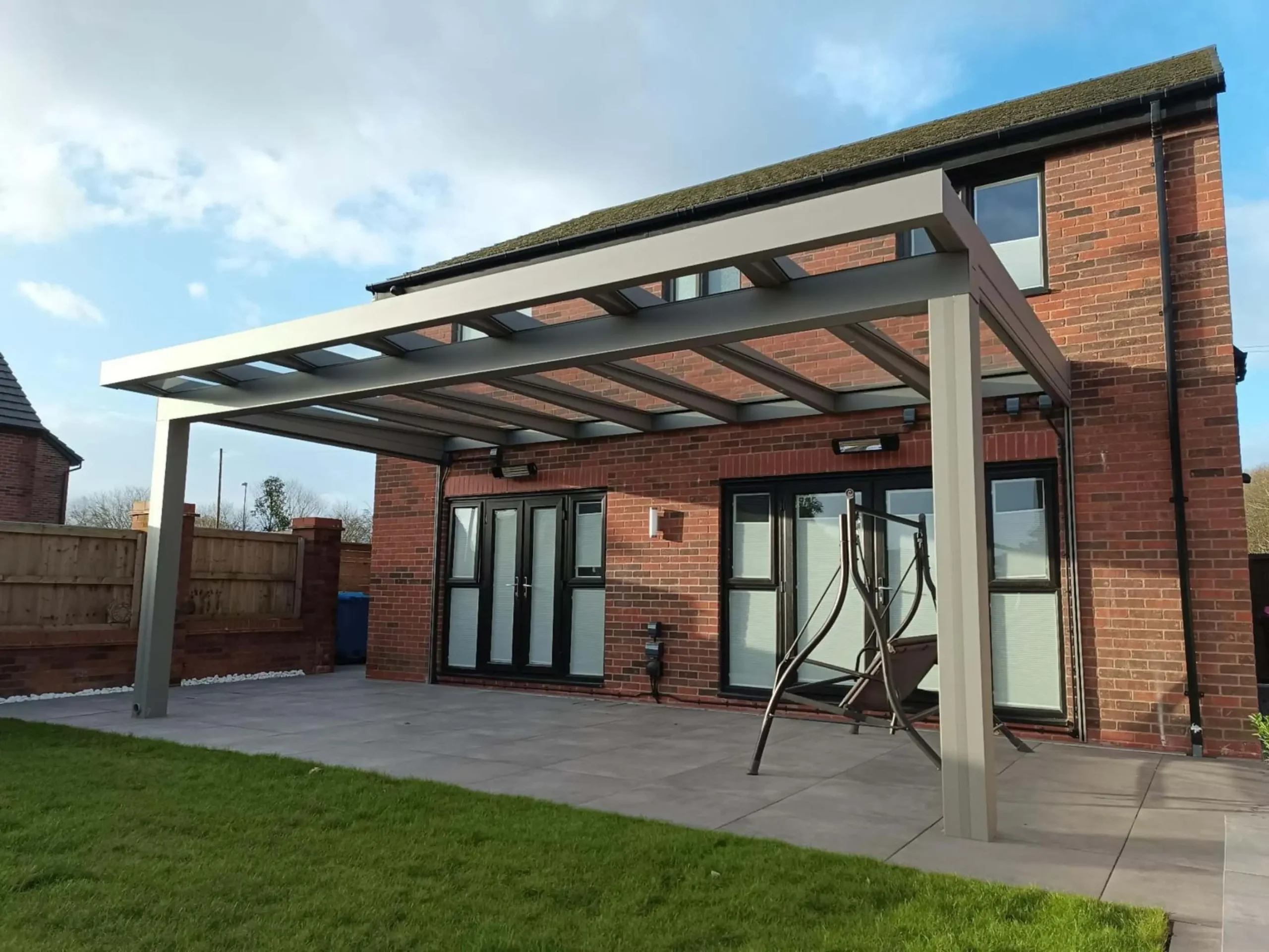 A silver grey aluminium cube line veranda with a laminated glass roof, remote-controlled side screens and black wall-mounted infrared heaters, furnished with a dark grey 3 seater swing chair.