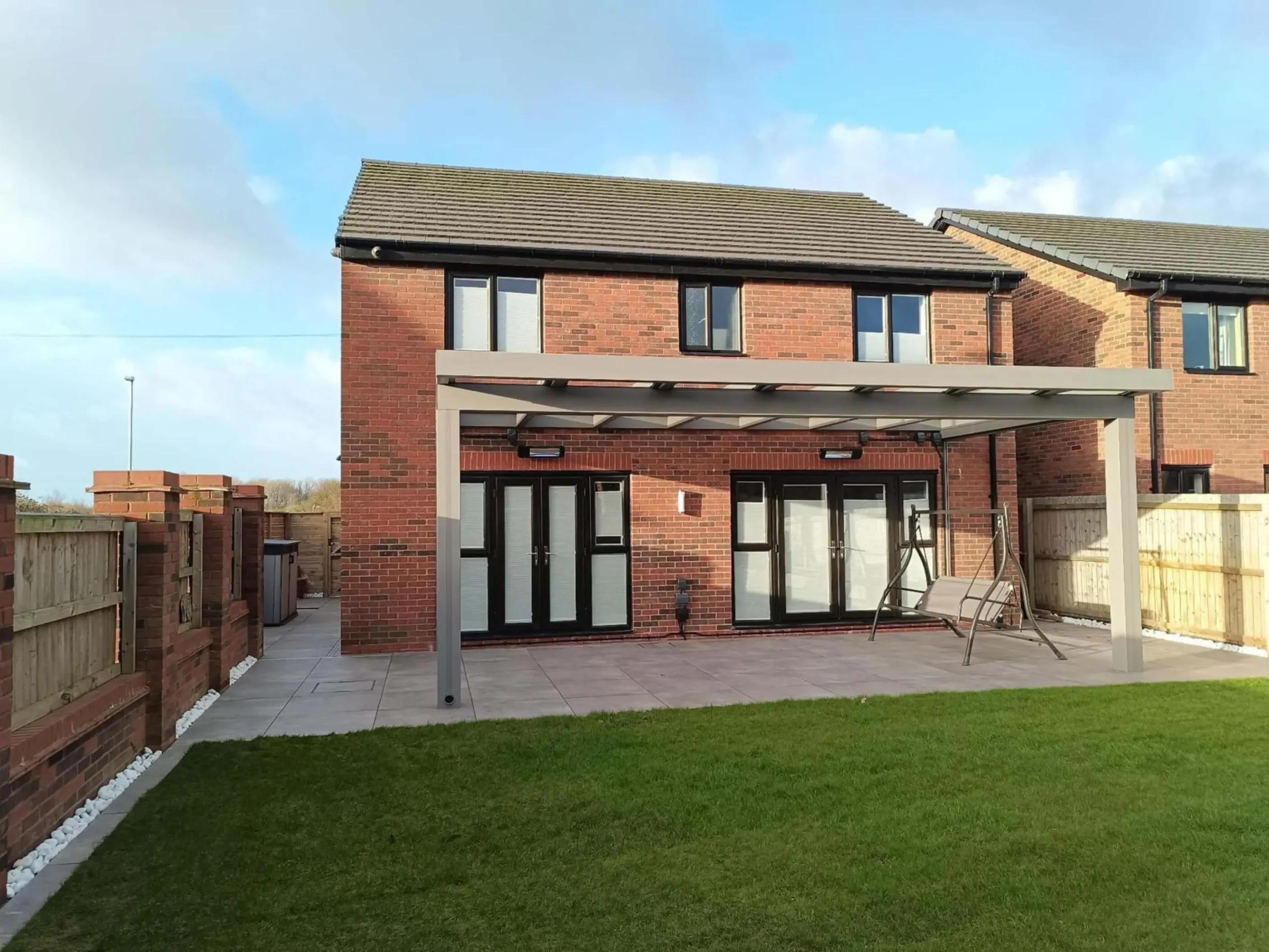 A silver grey aluminium cube line veranda with a laminated glass roof, remote-controlled side screens and black wall-mounted infrared heaters, furnished with a dark grey 3 seater swing chair.
