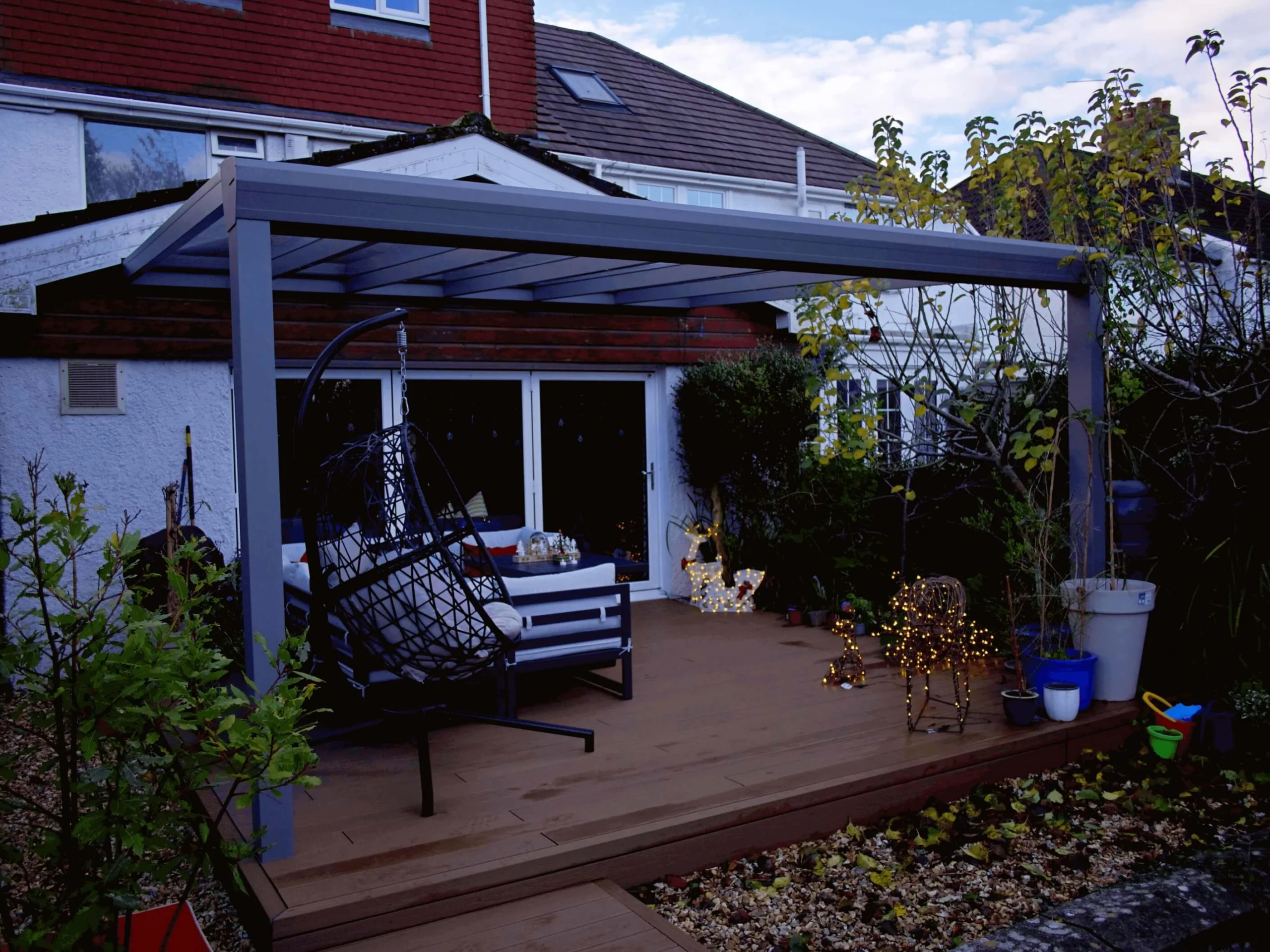 Anthracite grey glass roof veranda furnished with a black hammock chair, and Christmas decor and lighting, on a wooden deck.