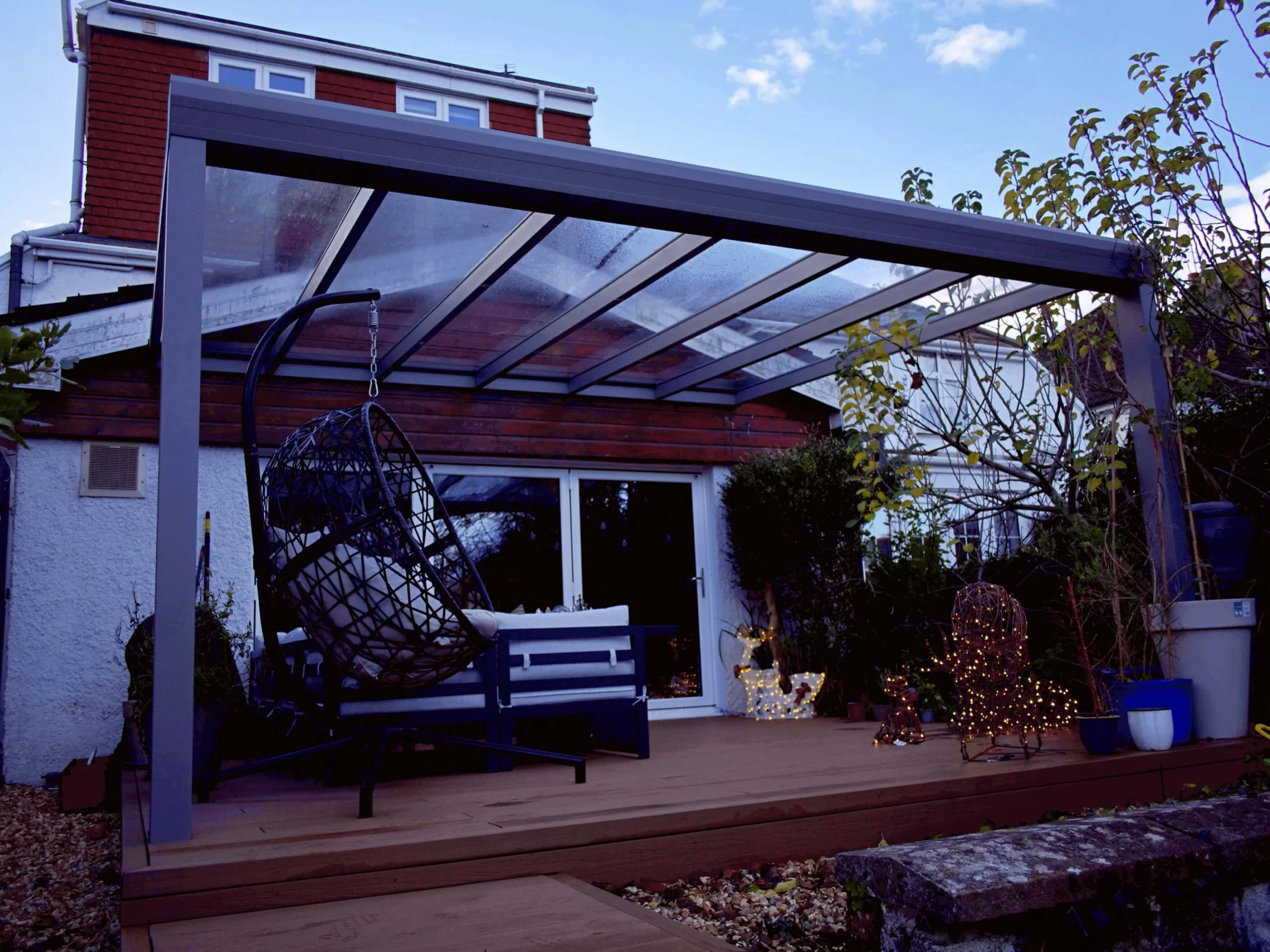 Anthracite grey glass roof veranda furnished with a black hammock chair, and Christmas decor and lighting, on a wooden deck.