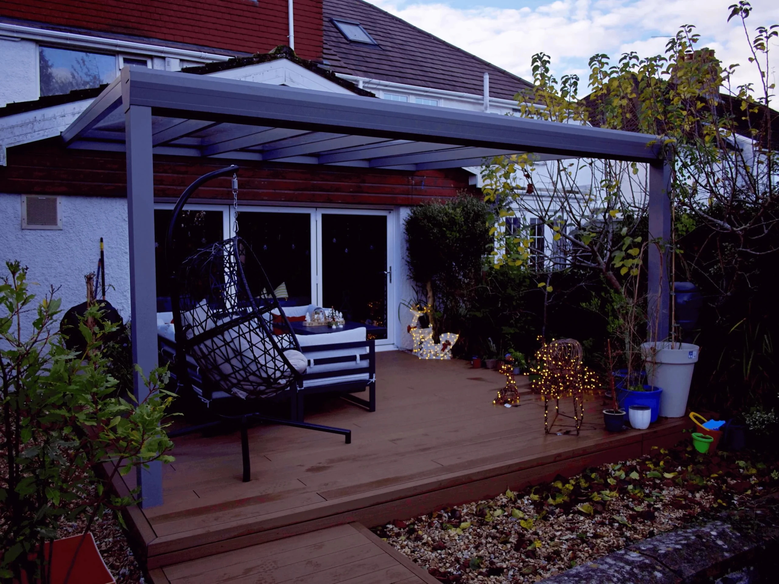 Anthracite grey glass roof veranda furnished with a black hammock chair and Christmas decor and lighting, on a wooden deck.