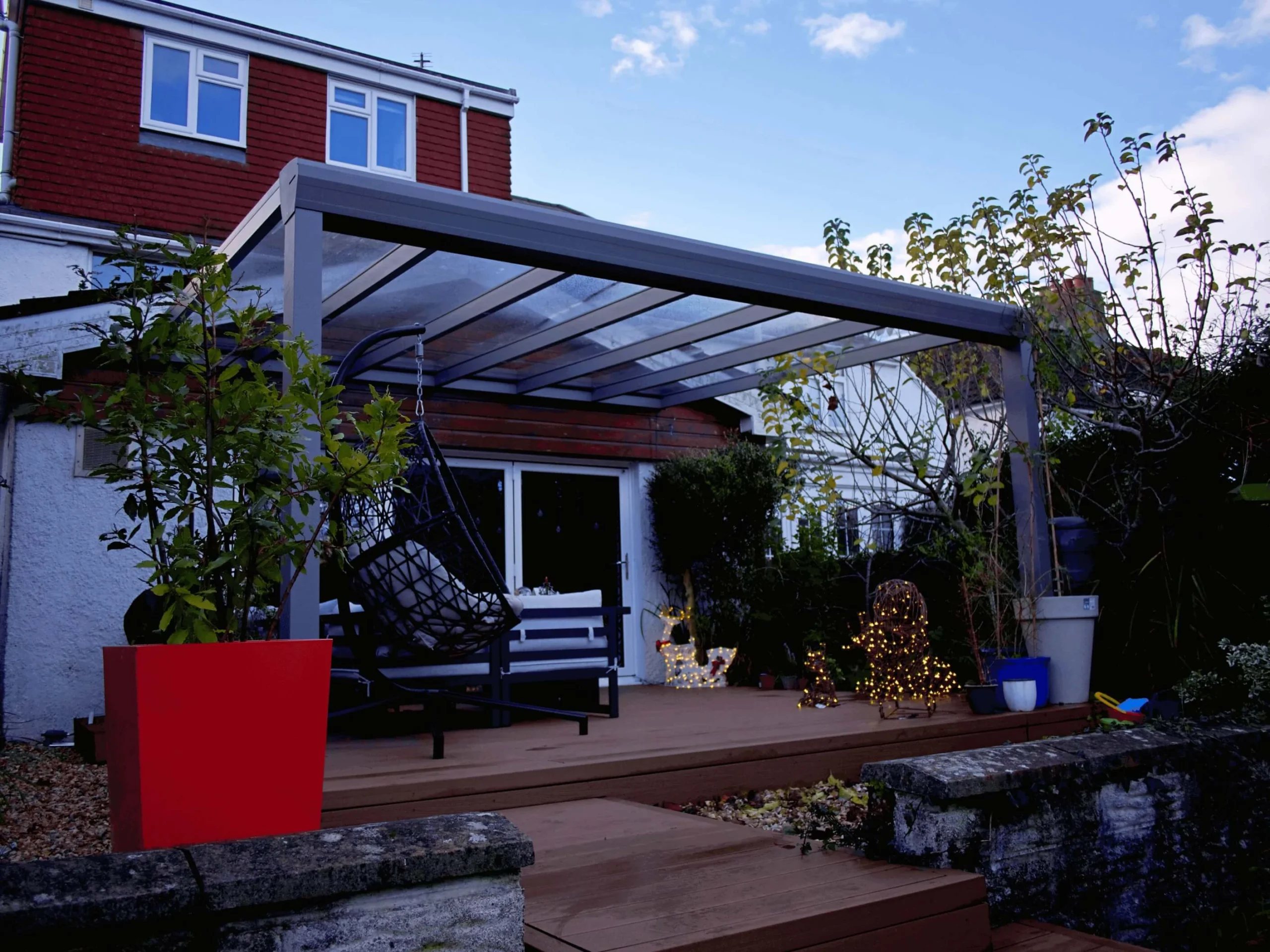 Anthracite grey glass roof veranda furnished with a black hammock chair, large red plant pot and Christmas decor and lighting, on a wooden deck.
