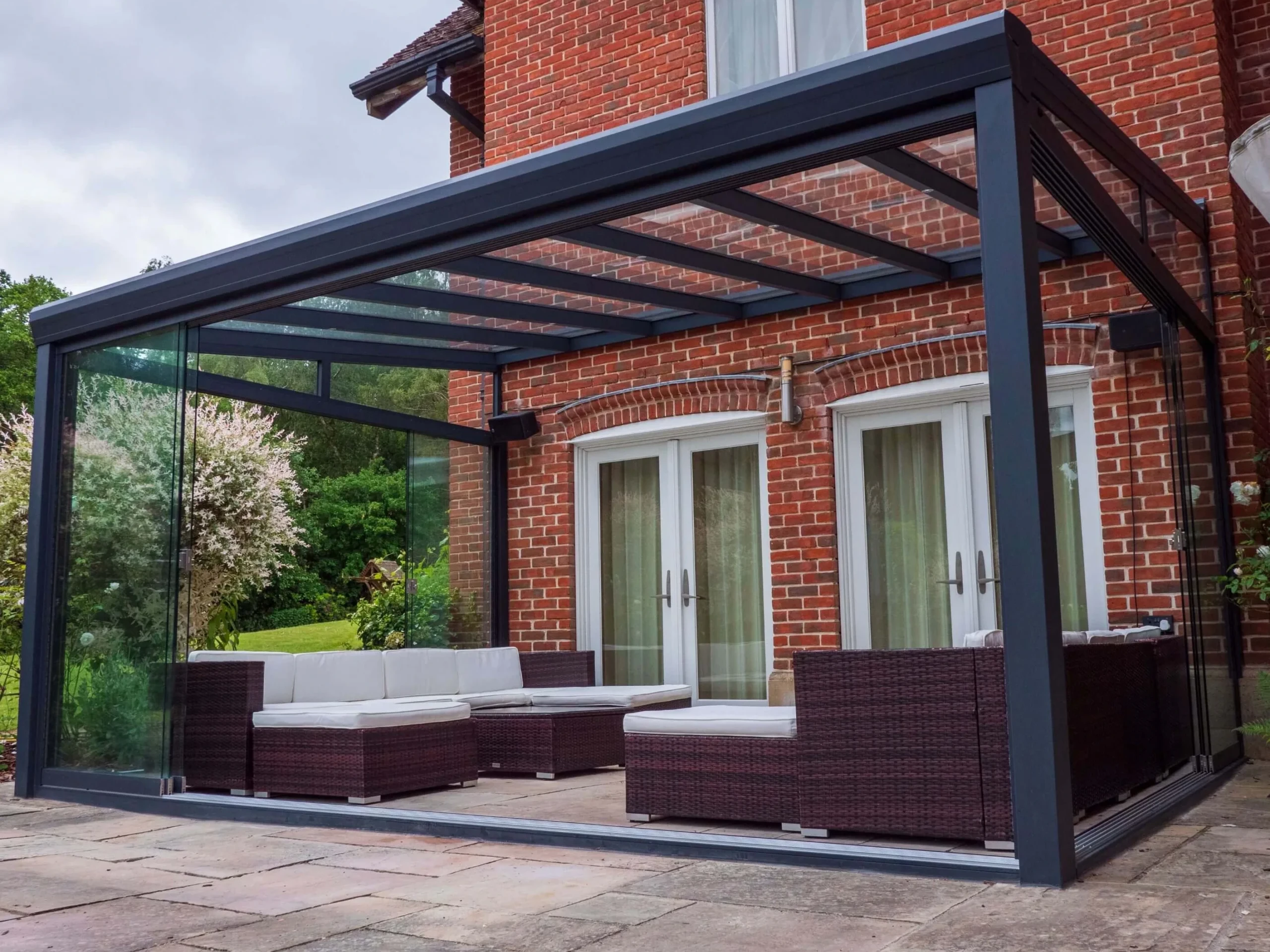 Anthracite grey aluminium frame glass room veranda with lockable sliding doors, furnished with brown rattan garden sofas, cream white cushions and black outdoor speakers.