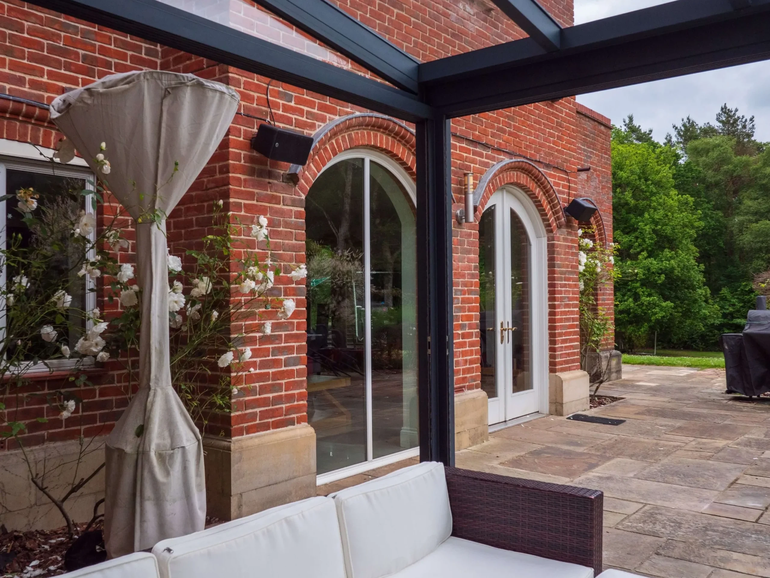 Anthracite grey aluminium frame glass room veranda with lockable sliding doors, furnished with brown rattan garden sofas, cream white cushions and black outdoor speakers.