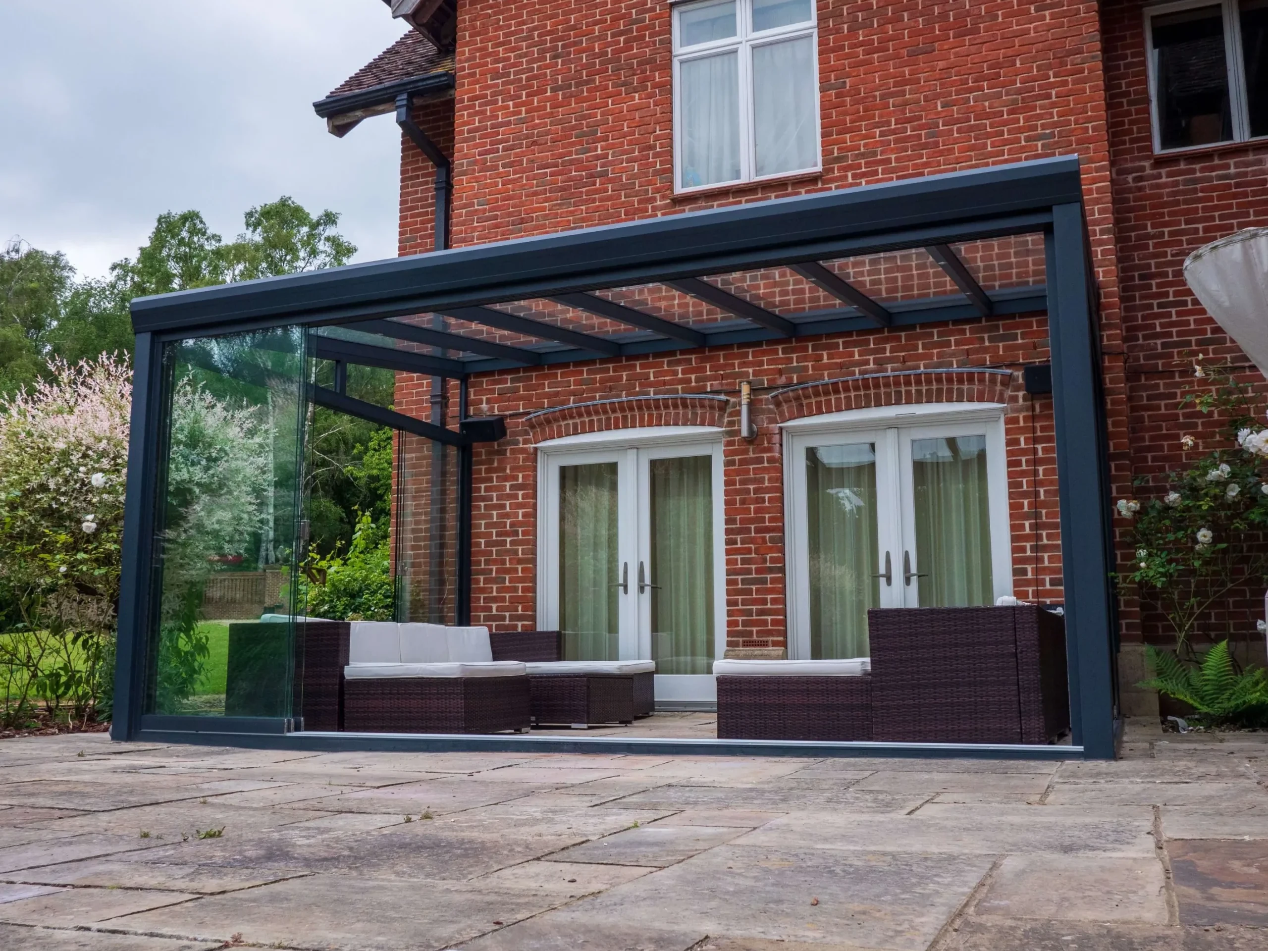 Anthracite grey aluminium frame glass room veranda with lockable sliding doors, furnished with brown rattan garden sofas, cream white cushions and black outdoor speakers.