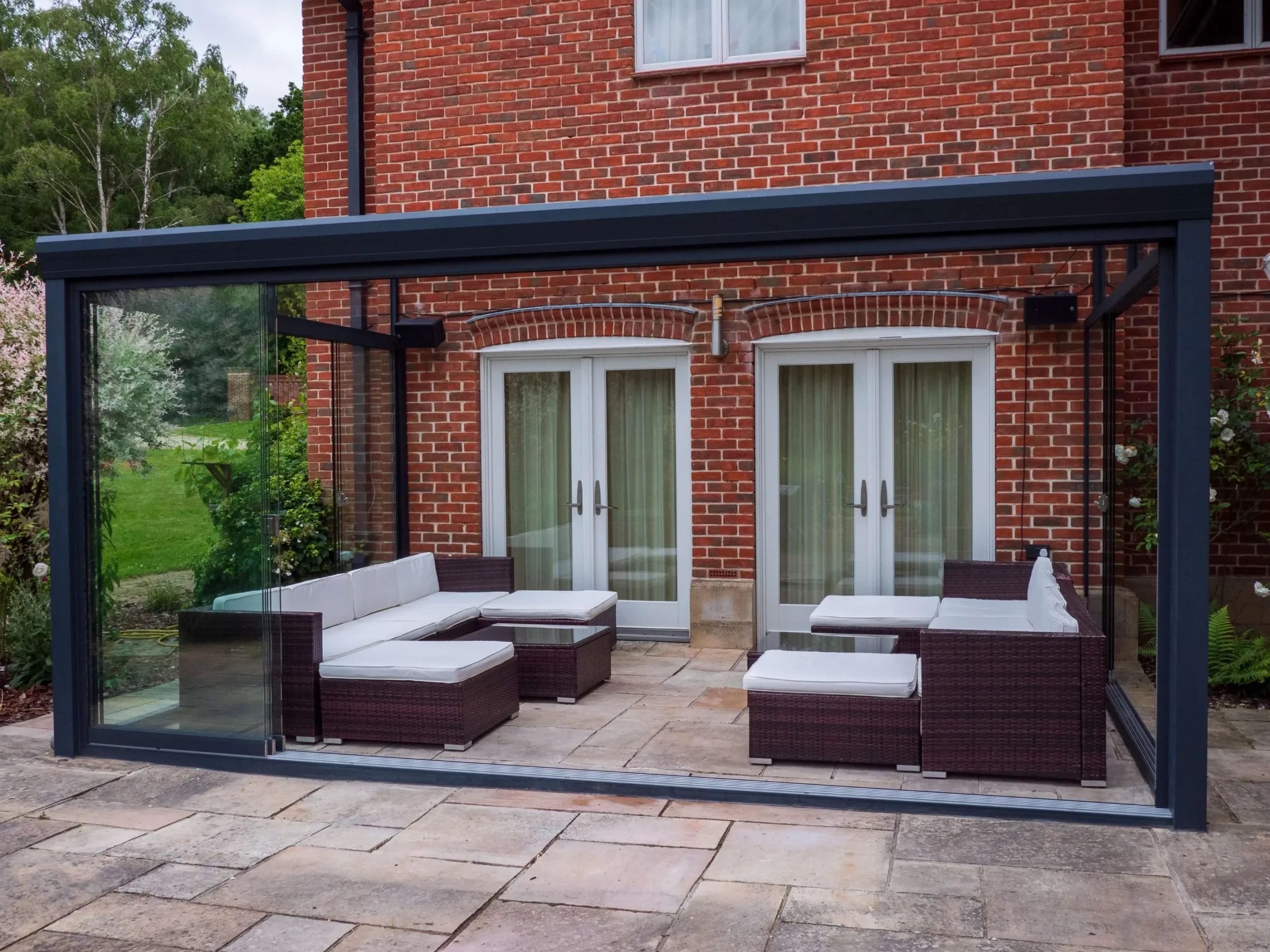 Anthracite grey aluminium frame glass room veranda with lockable sliding doors, furnished with brown rattan garden sofas, cream white cushions and black outdoor speakers.