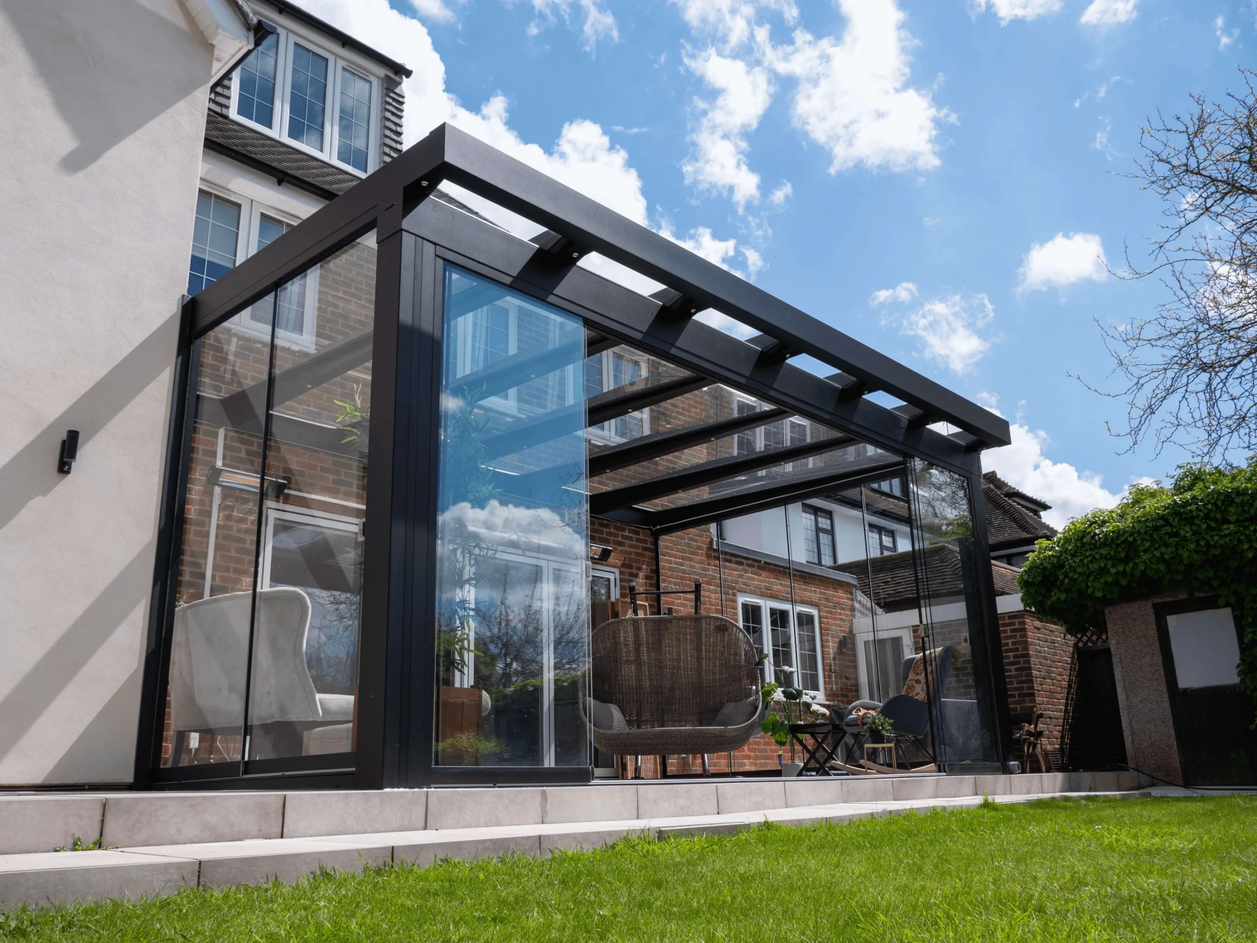 Jet black frame aluminium glass room veranda with an overhang roof, fixed glass walls and glass sliding doors which are opened to showcase the brown hammock chair in the interior.