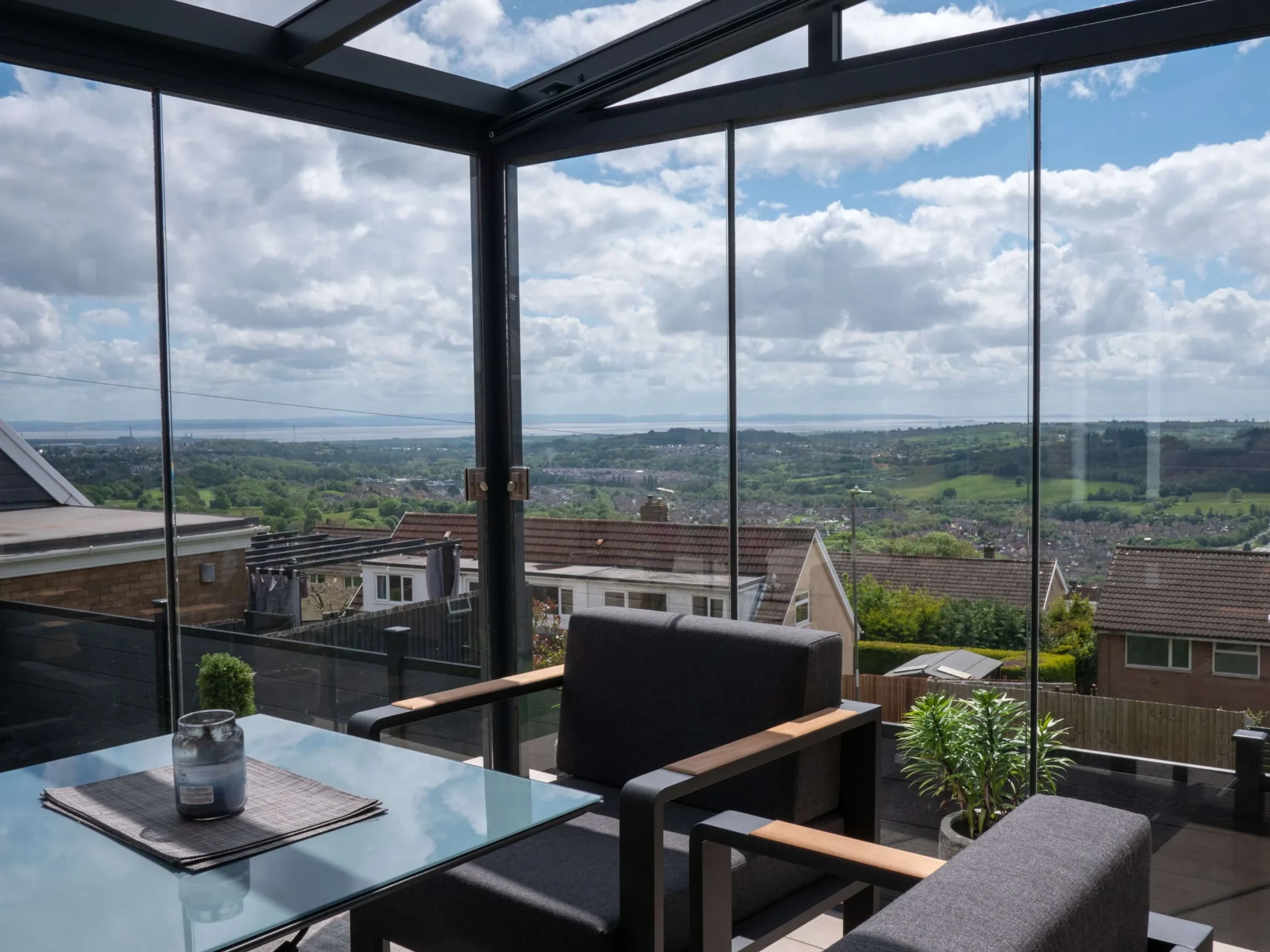 Interior of an anthracite grey aluminium framed glass room veranda with lockable glass sliding doors and LED spot lights, furnished with a glass dining table and grey sofa chairs.