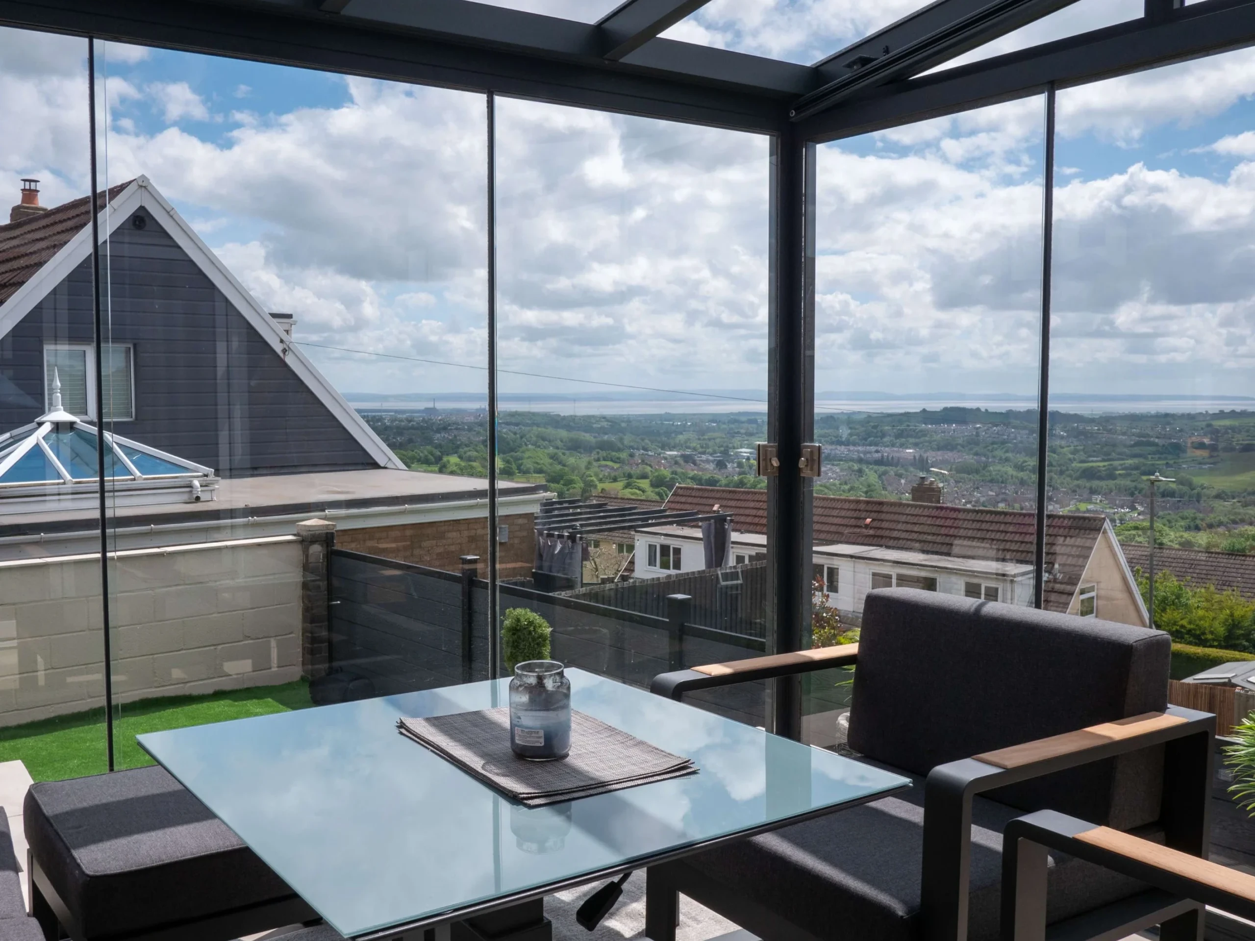 Interior of an anthracite grey aluminium framed glass room veranda with lockable glass sliding doors and LED spot lights, furnished with a glass dining table and grey sofa chairs.