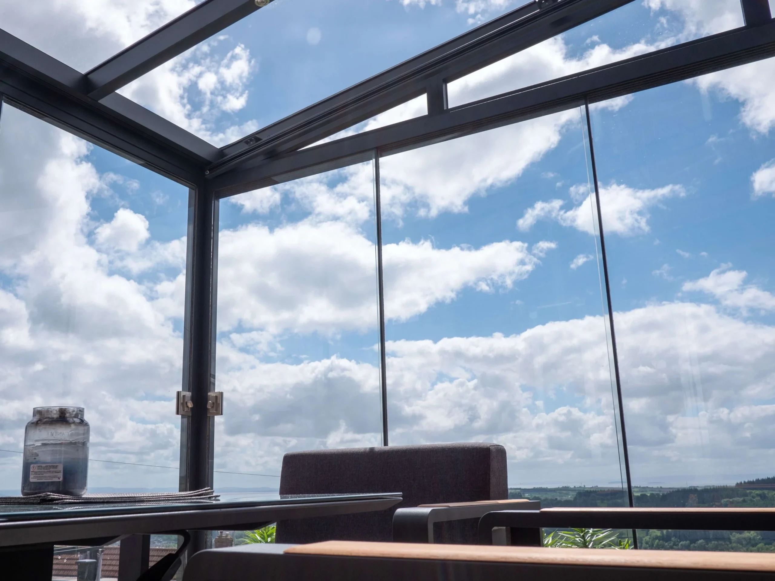 Interior of an anthracite grey aluminium framed glass room veranda with lockable glass sliding doors and LED spot lights, furnished with a glass dining table and grey sofa chairs.