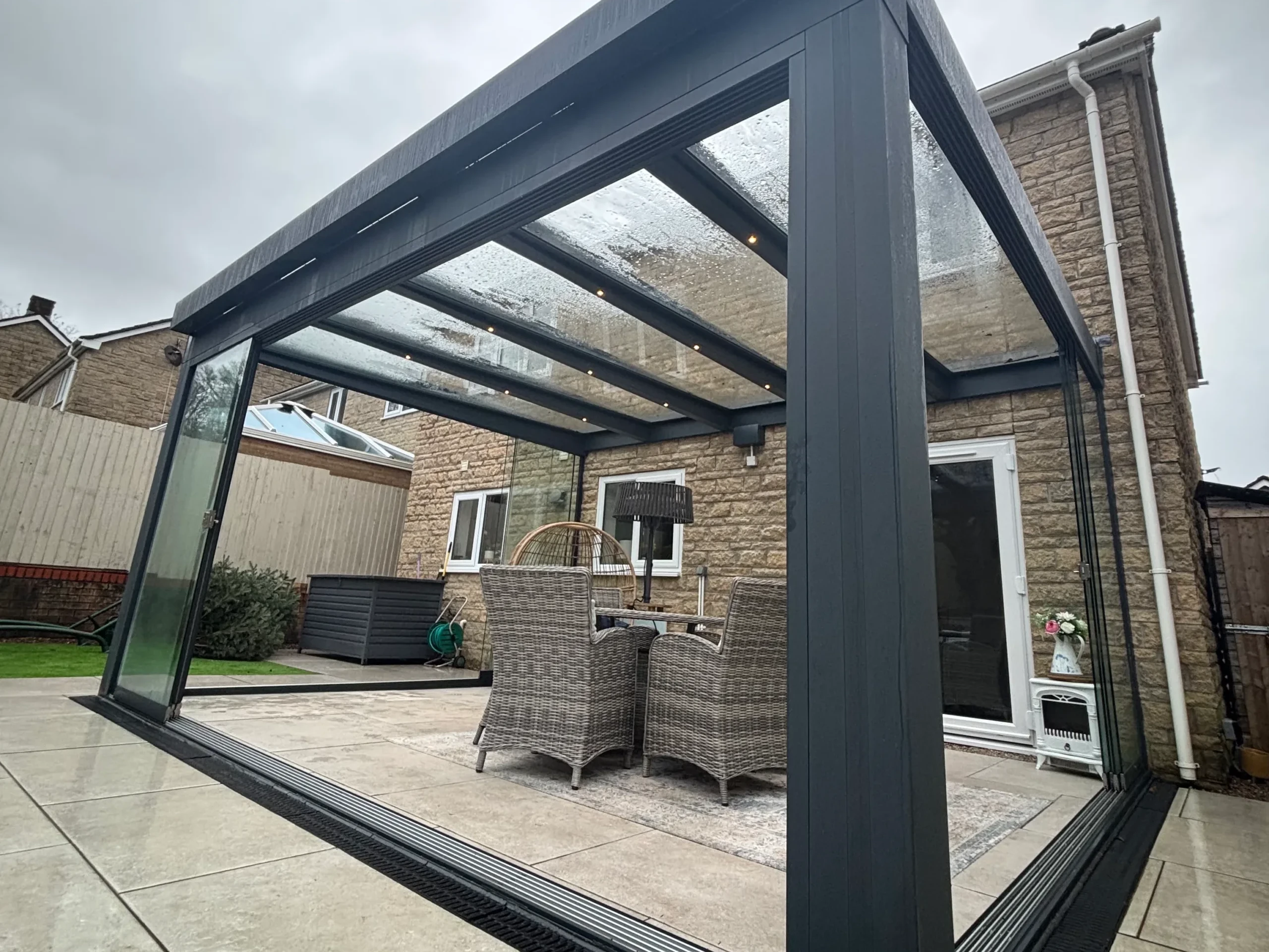 Anthracite grey aluminium framed glass room veranda with opened glass sliding doors, furnished with a beige rattan garden dining set and brown hammock chairs.