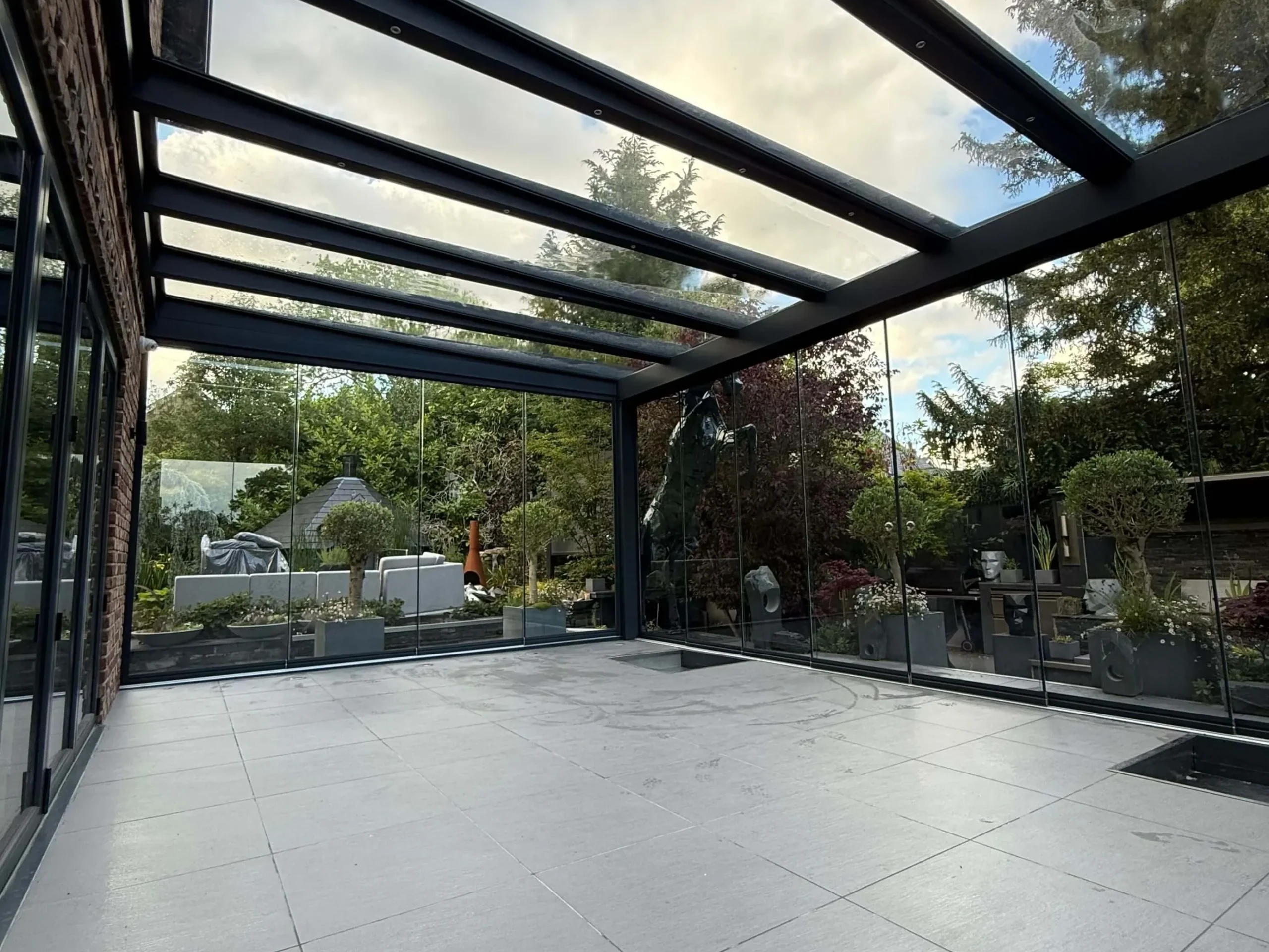 Anthracite grey frame glass room veranda on a cream white porcelain patio with a clear glass roof, lockable sliding doors and silver spot lights.