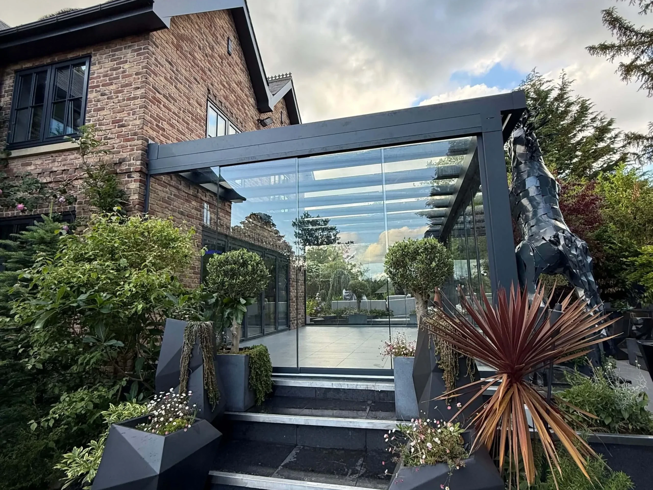 Anthracite grey frame glass room veranda on a cream white porcelain patio with a clear glass roof and lockable sliding doors, externally decorated with a black prancing horse statue, futuristic outdoor plant pots and piano black porcelain steps.