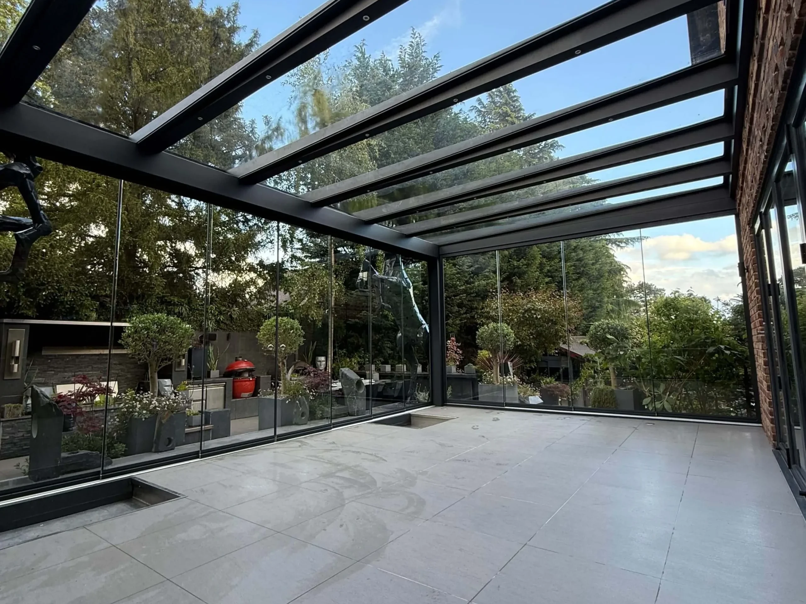 Anthracite grey frame glass room veranda on a cream white porcelain patio with a clear glass roof, lockable sliding doors and silver spot lights.