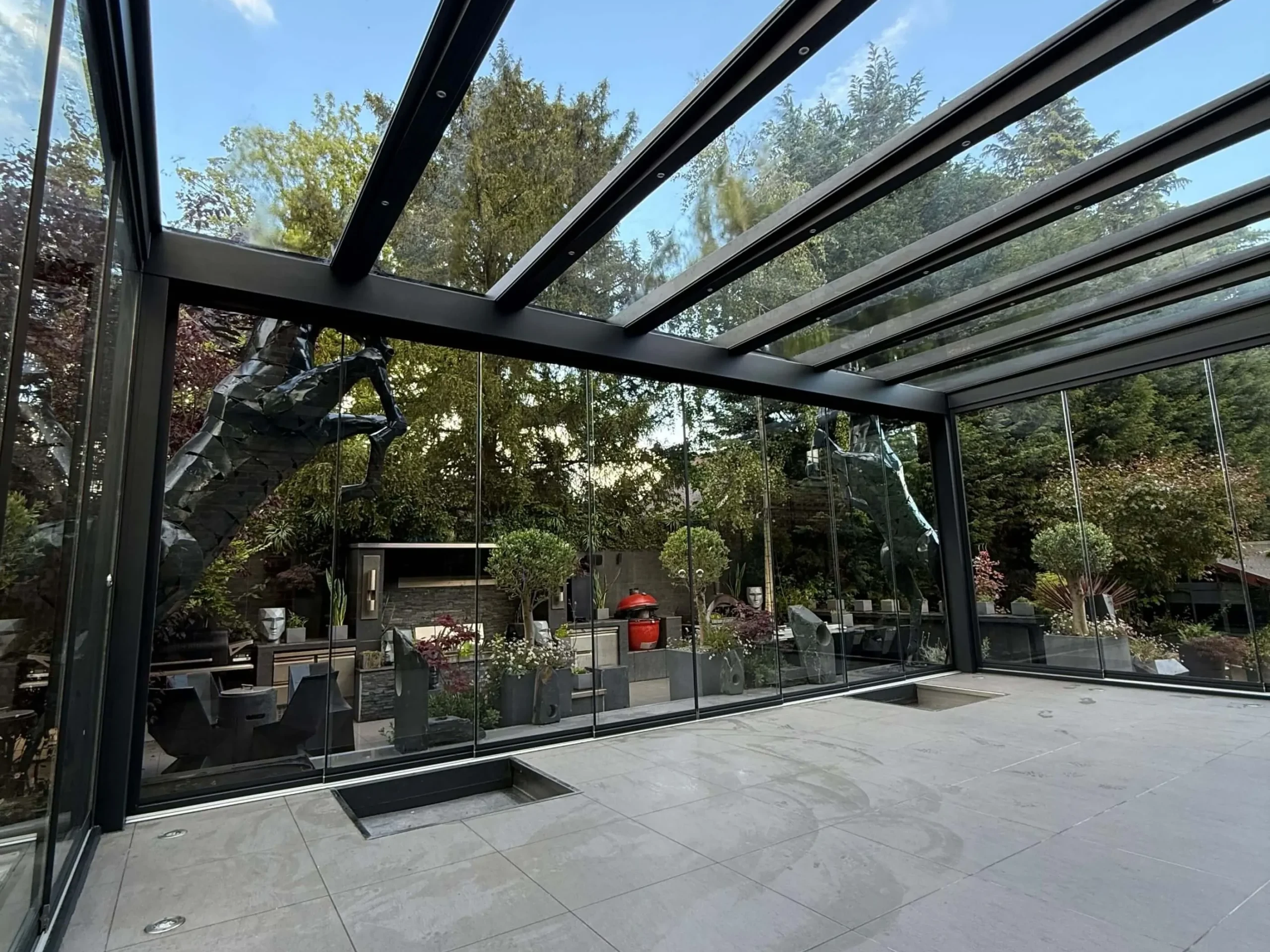 Anthracite grey frame glass room veranda on a cream white porcelain patio with a clear glass roof, lockable sliding doors and silver spot lights.
