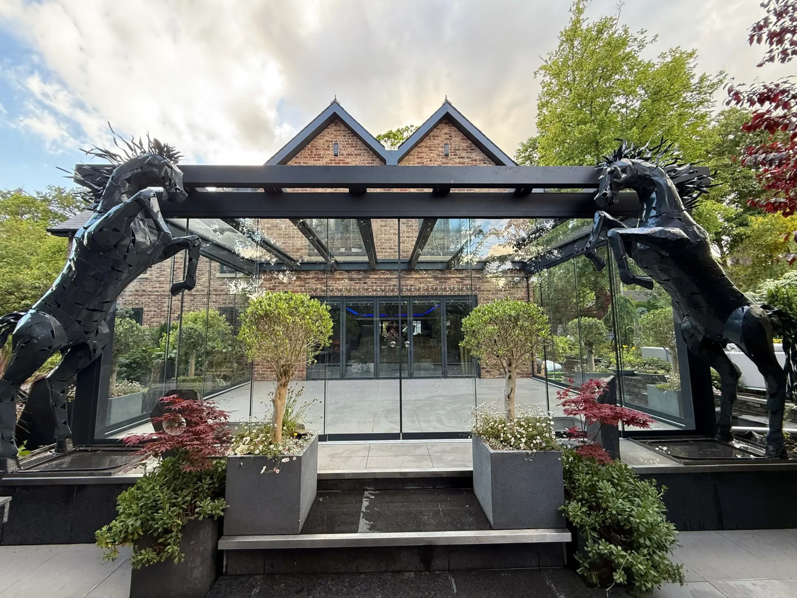 Anthracite grey frame glass room veranda on a cream white porcelain patio with a clear glass roof and lockable sliding doors, externally decorated with black prancing horse statues and a mixture of outdoor plants.
