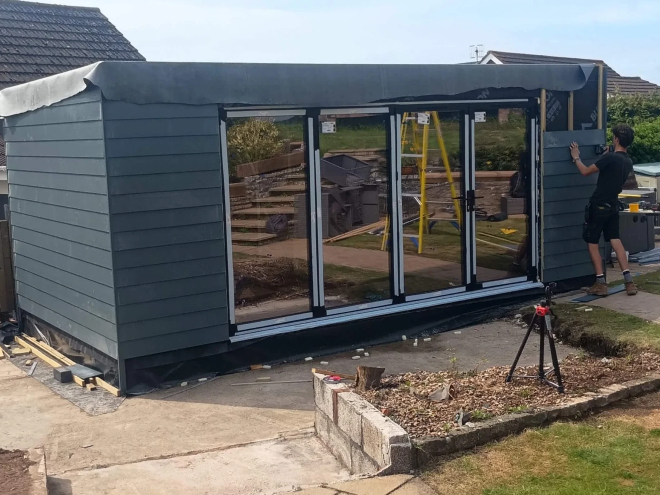 Unfinished grey composite cladded insulated garden room with black aluminium framed bifold doors and a black rubber roof.
