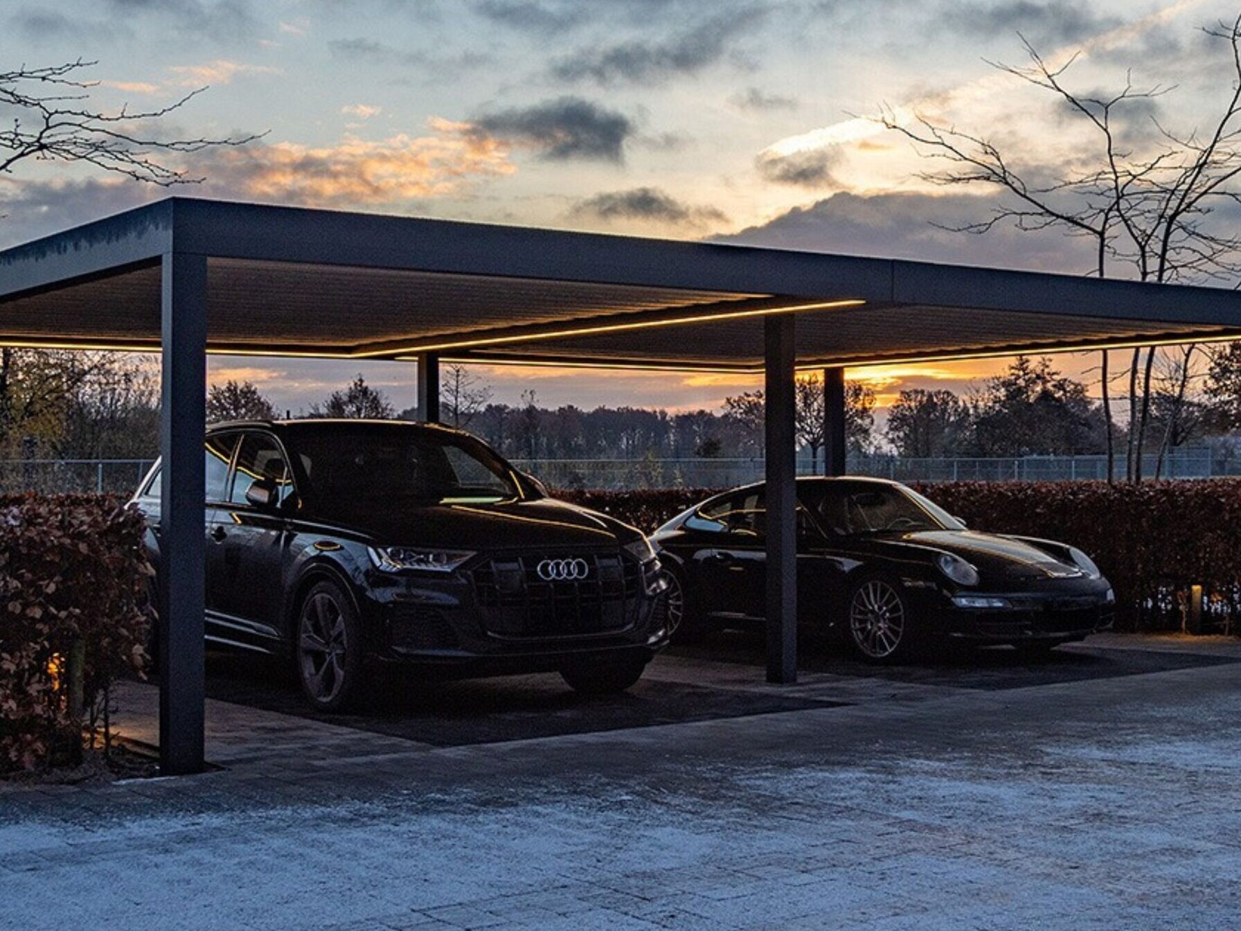 Anthracite grey aluminium frame double carport with LED strip lighting in the evening, sheltering two black vehicle, an SUV and a sports car.