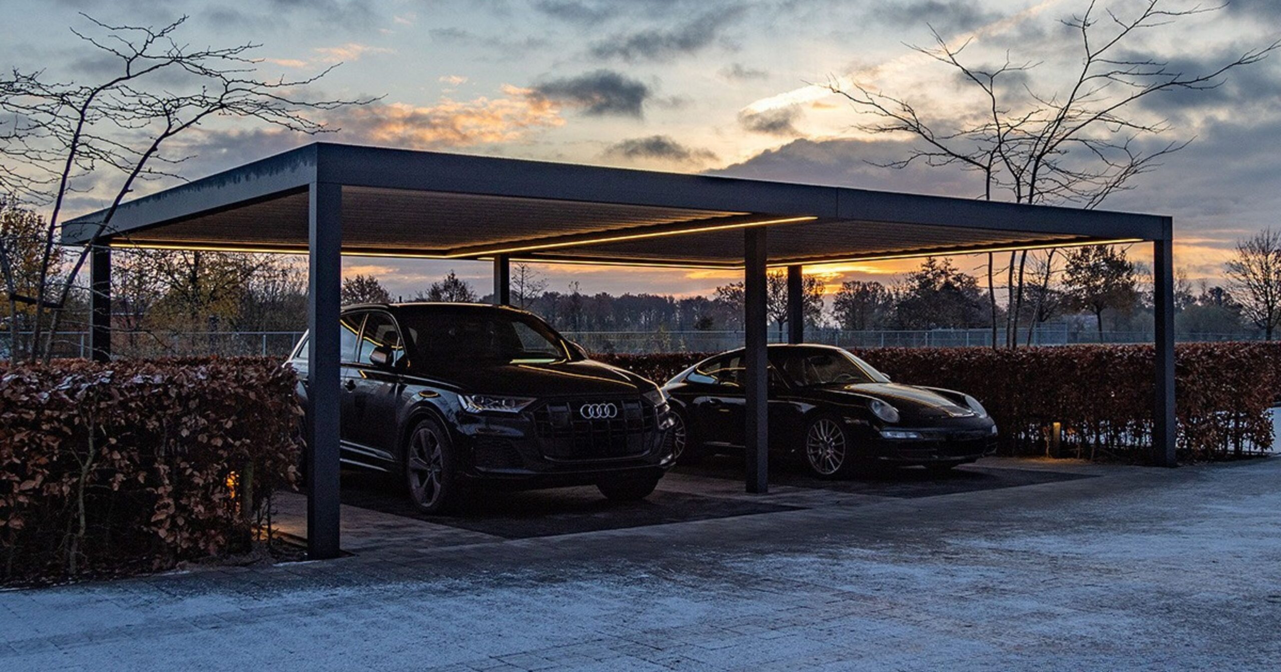 Anthracite grey aluminium frame double carport with LED strip lighting in the evening, sheltering two black vehicle, an SUV and a sports car.