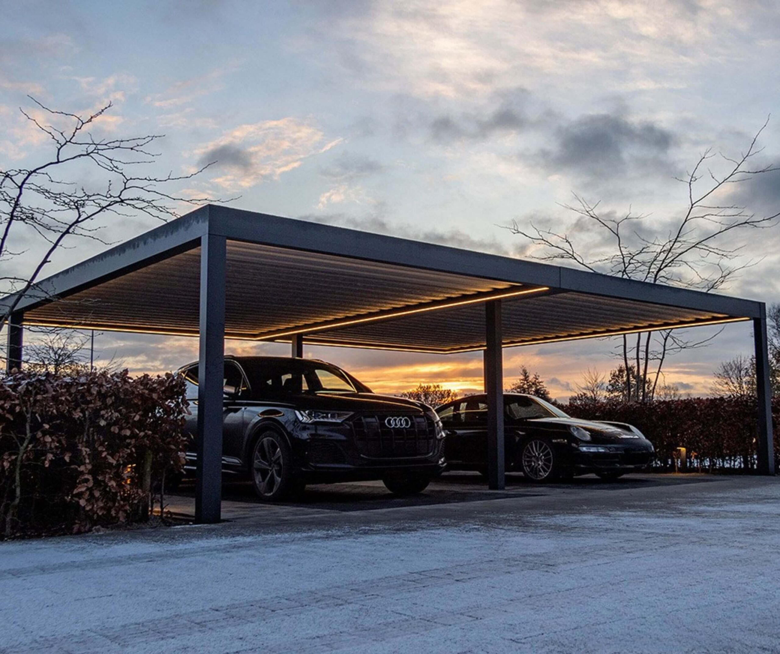 Anthracite grey aluminium frame double carport with LED strip lighting in the evening, sheltering two black vehicle, an SUV and a sports car.