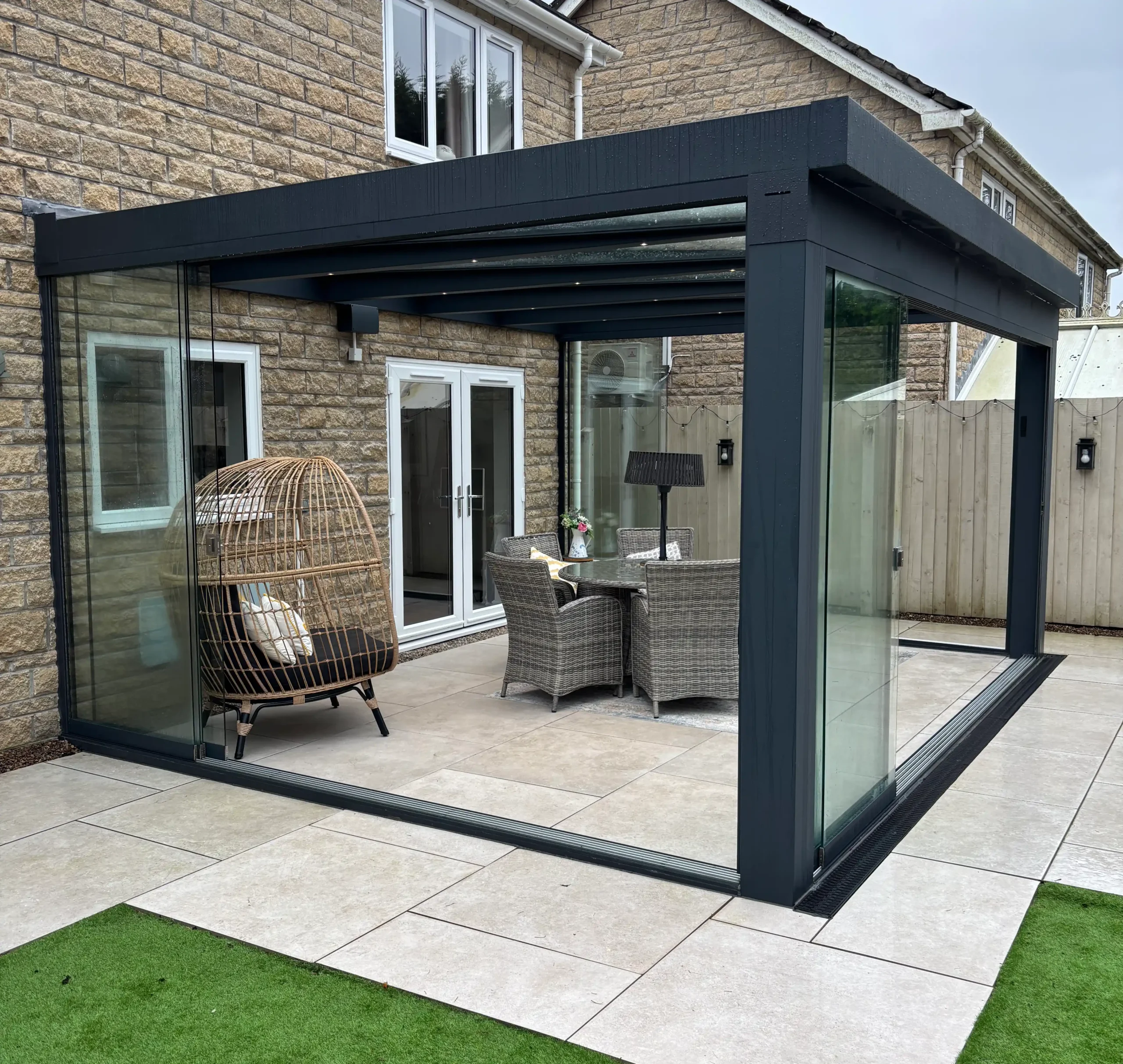 Anthracite grey aluminium framed glass room veranda with opened glass sliding doors, furnished with a beige rattan garden dining set and brown hammock chairs.