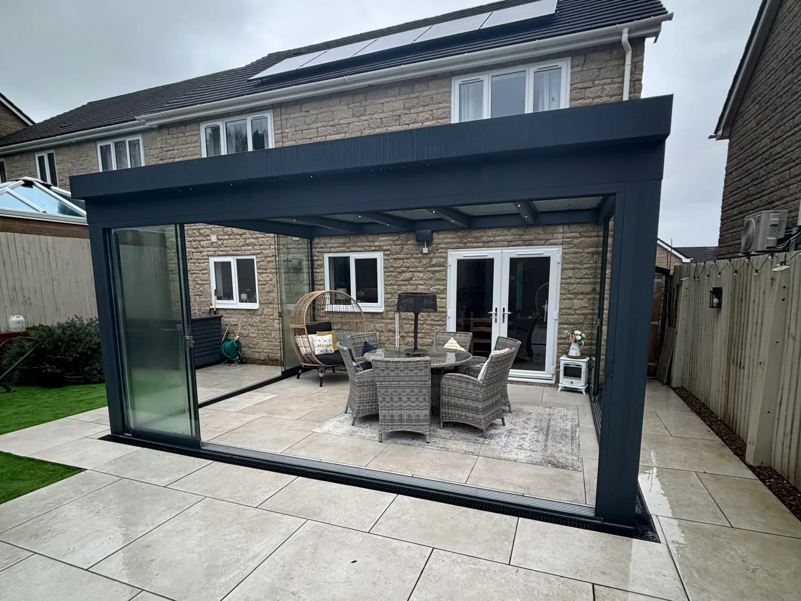 Anthracite grey aluminium framed glass room veranda with opened glass sliding doors, furnished with a beige rattan garden dining set and brown hammock chairs.